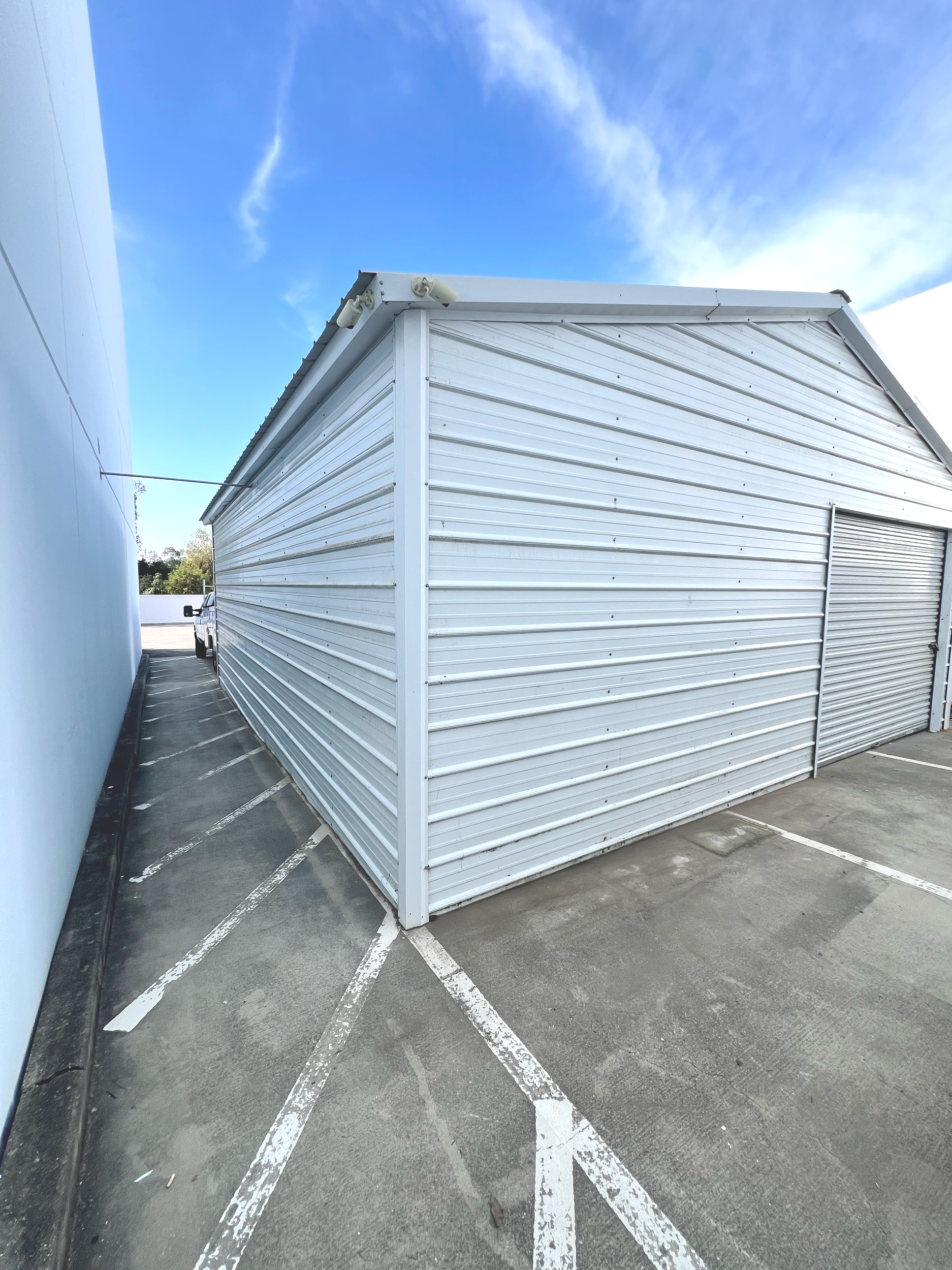 White metal storage shed with closed rolling door, situated in a parking lot with marked parking spaces, adjacent to a white wall, under a blue sky with some clouds.