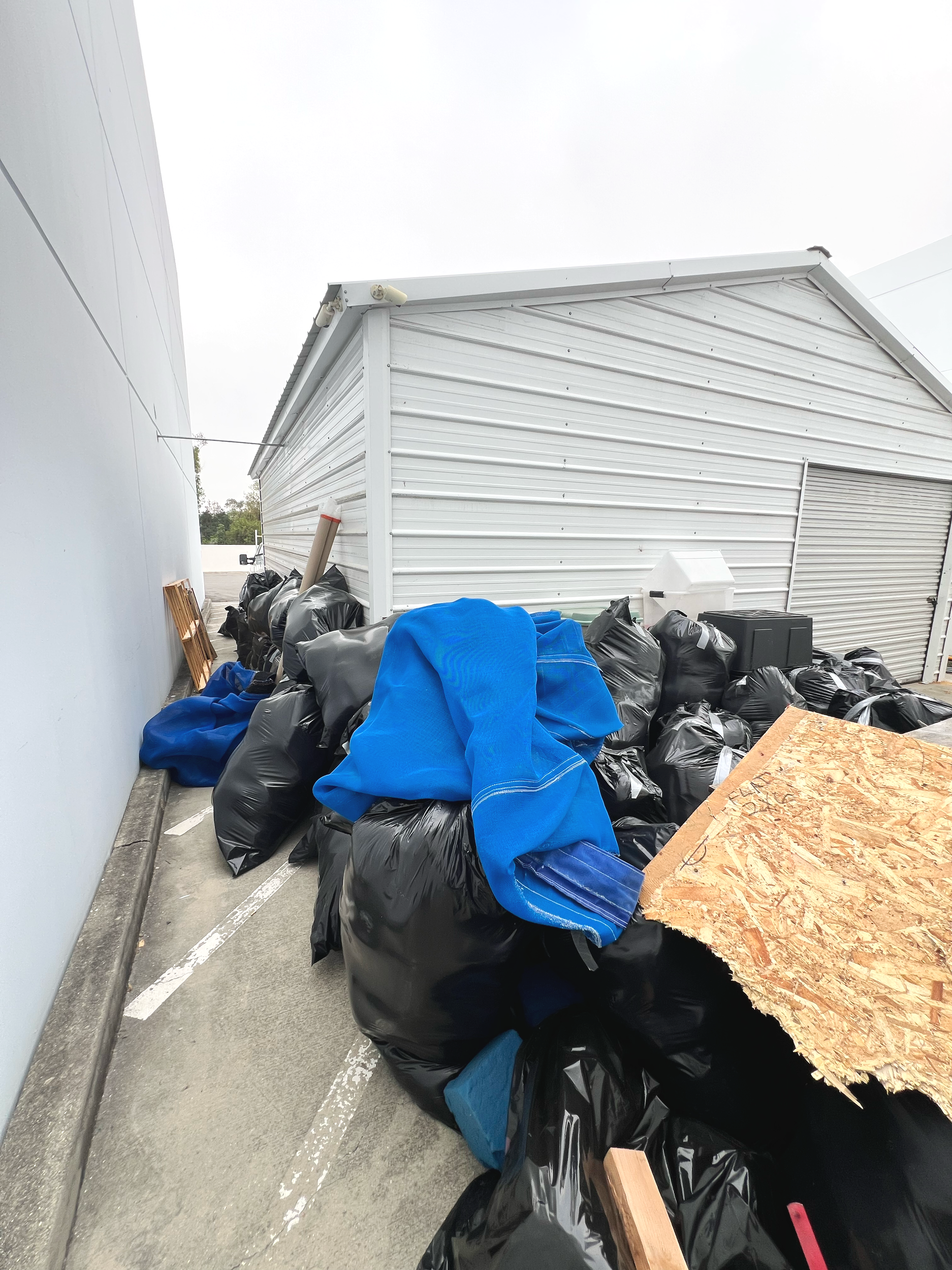 Piles of black trash bags, some with blue tarps, situated next to a white building with a metal garage door, on a concrete parking lot.