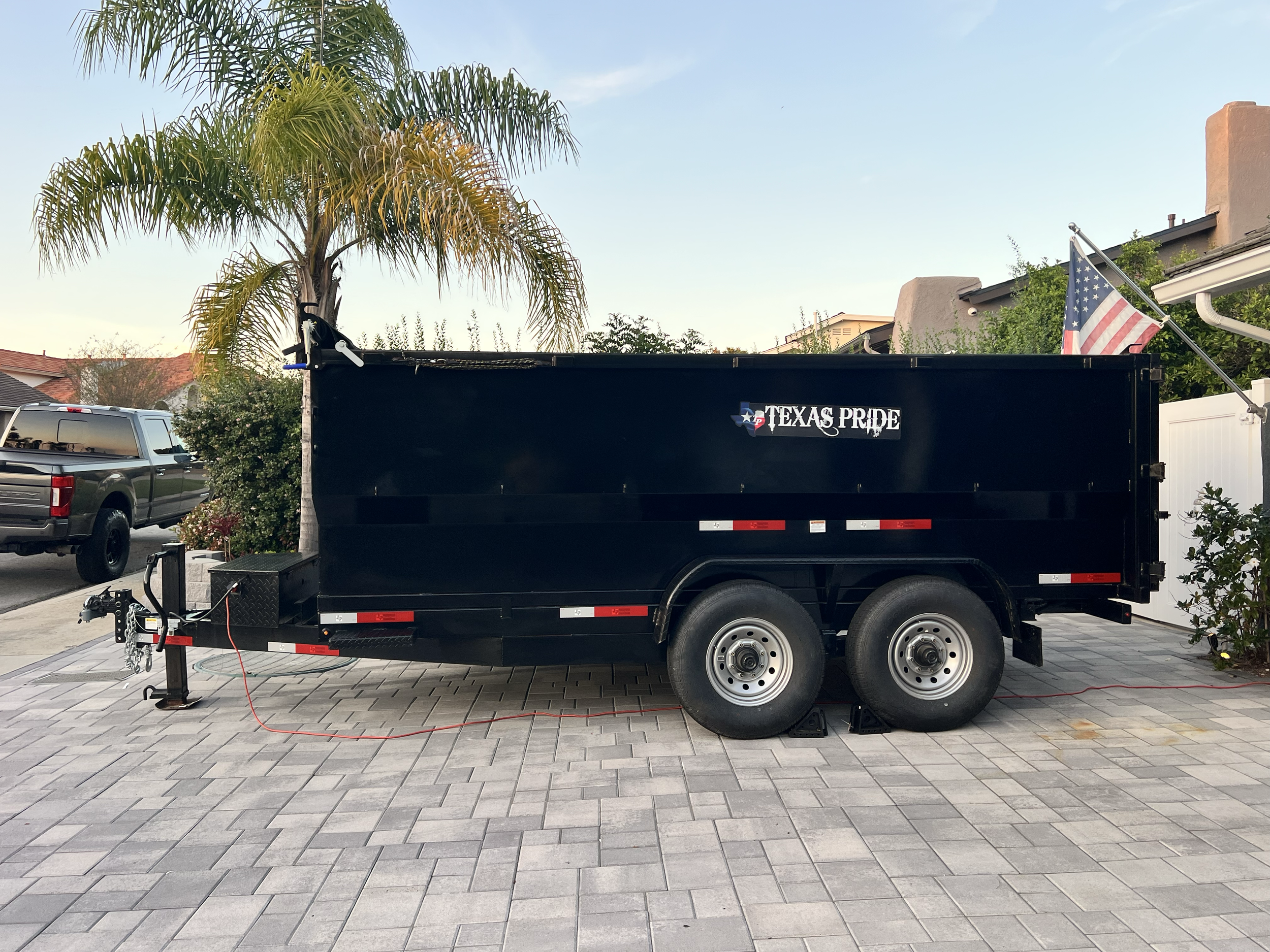 Black dump trailer with Texas Pride logo, parked on a paved driveway with a palm tree, a flag, and residential houses in the background.