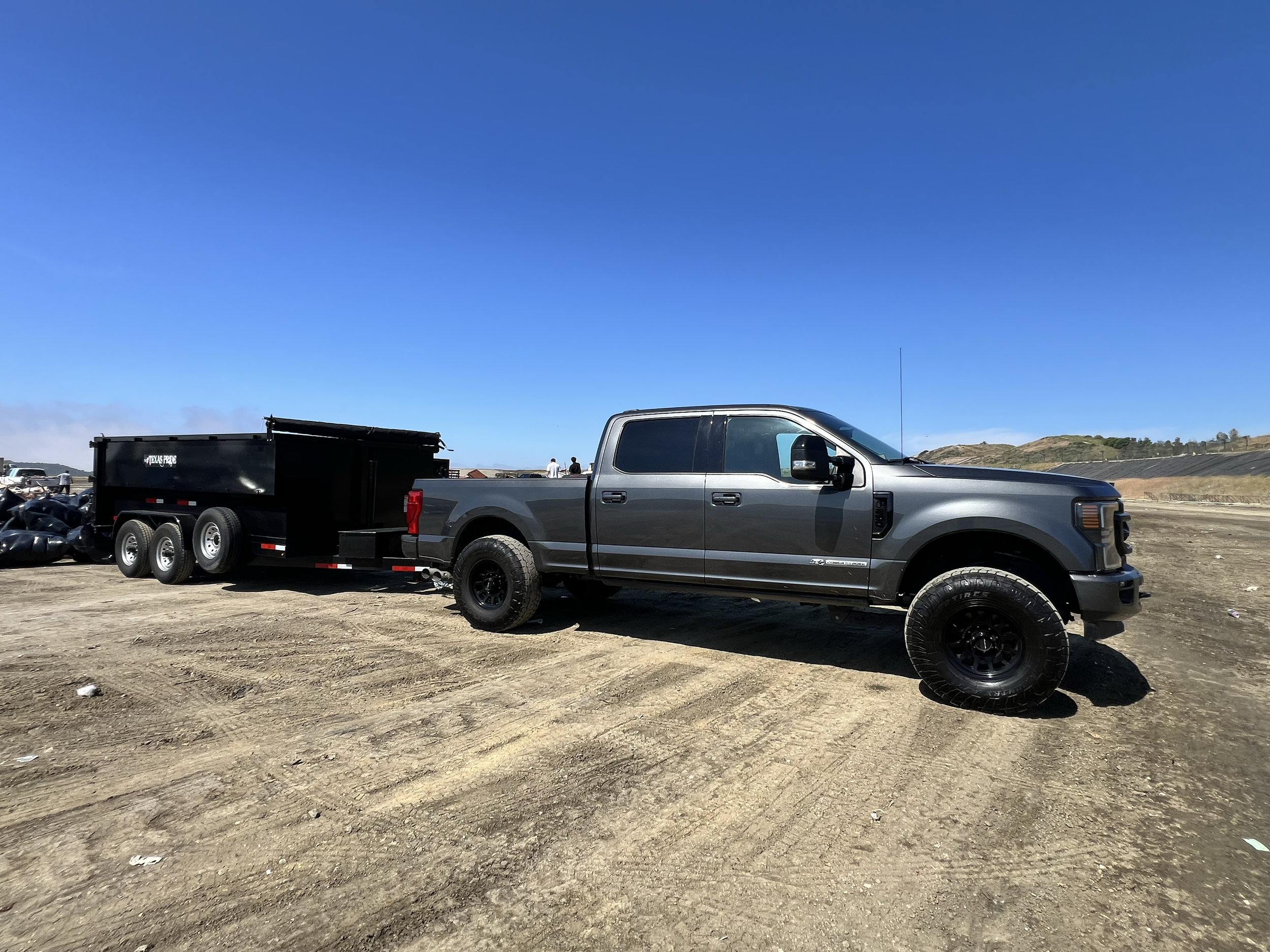 Gray pickup truck with a black trailer attached, parked on a dirt lot on a sunny day with a clear blue sky.
