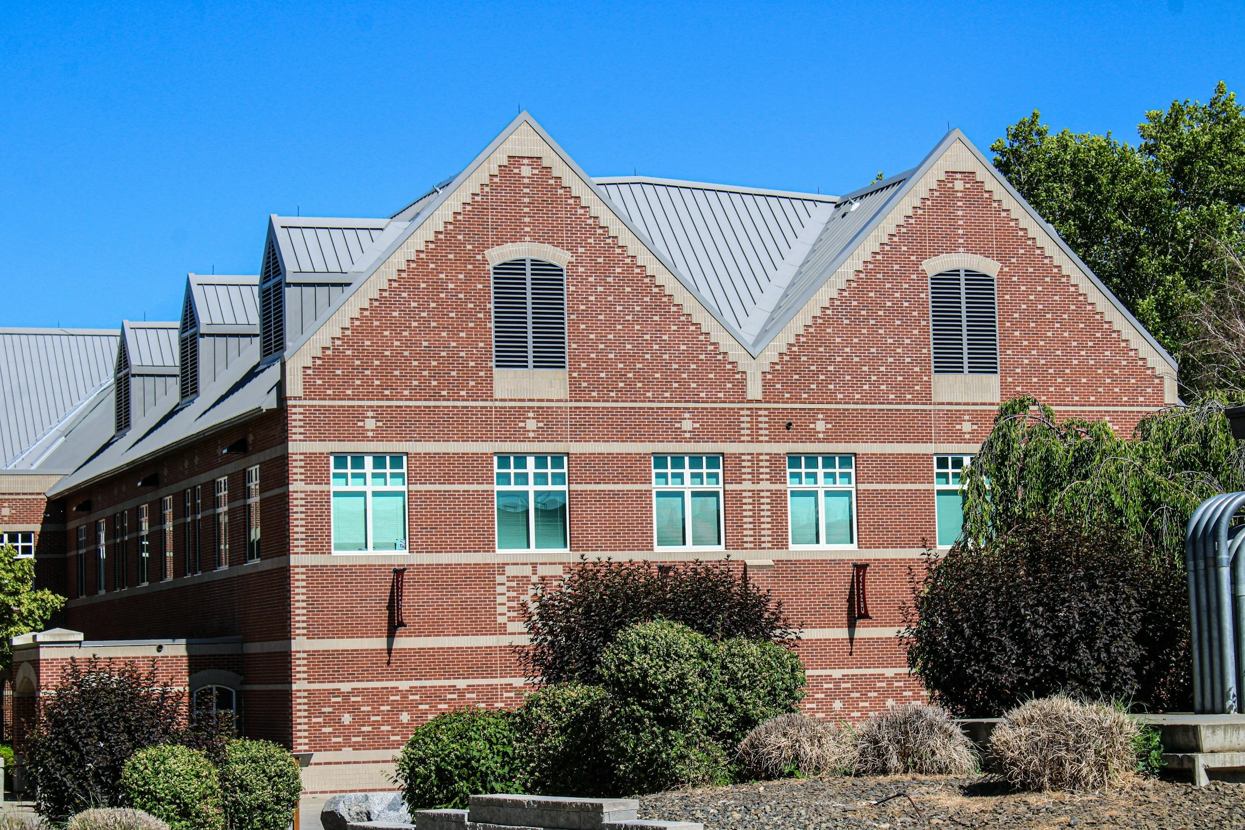 Red brick institutional building with decorative gabled roofs, tall windows, landscaped shrubs, and a bright blue sky