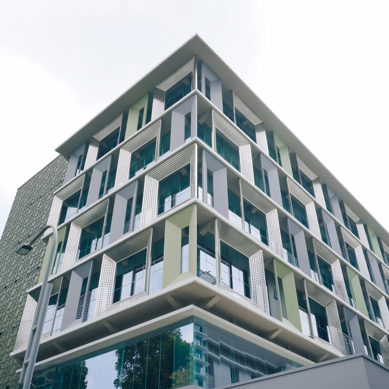 Modern multi-story office building with glass windows, geometric white facades, and perforated sunshade panels viewed from below