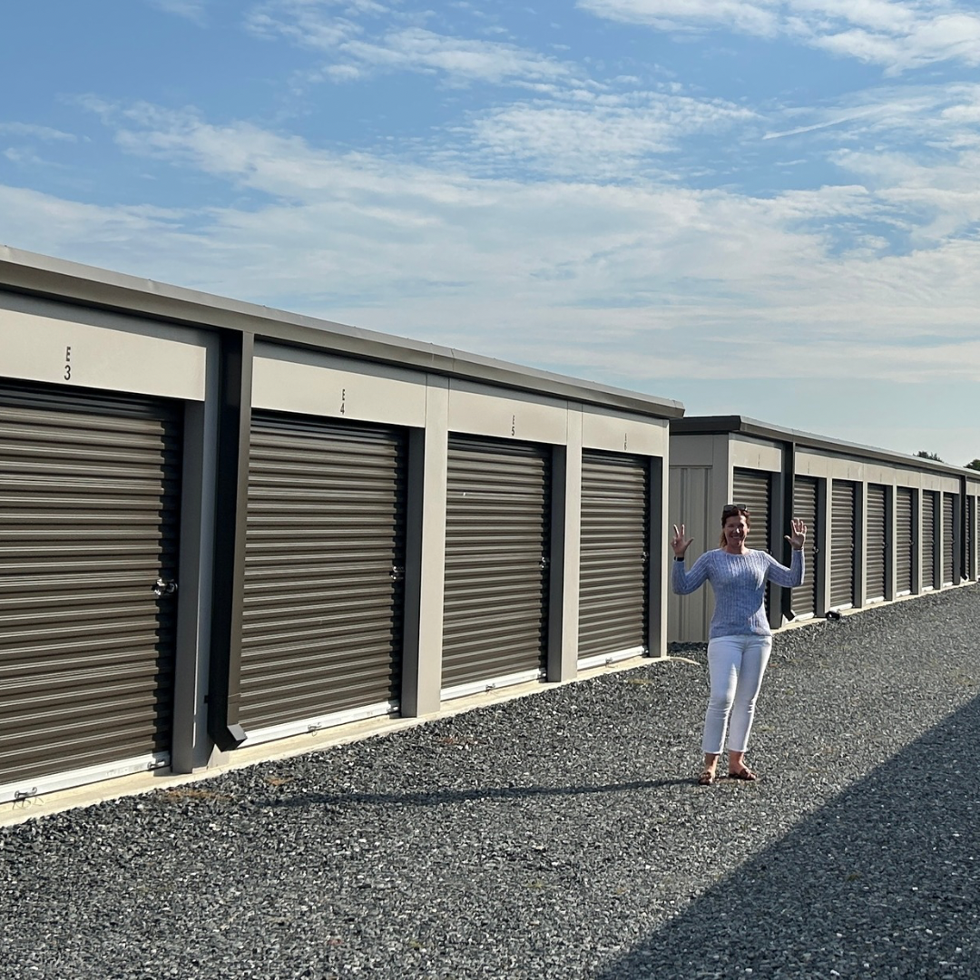 Woman standing with raised hands beside a row of gray storage unit doors under a blue sky