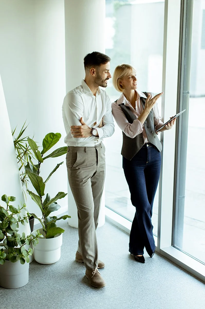 Business colleagues discussing a project beside office windows, with a woman holding a tablet and gesturing.