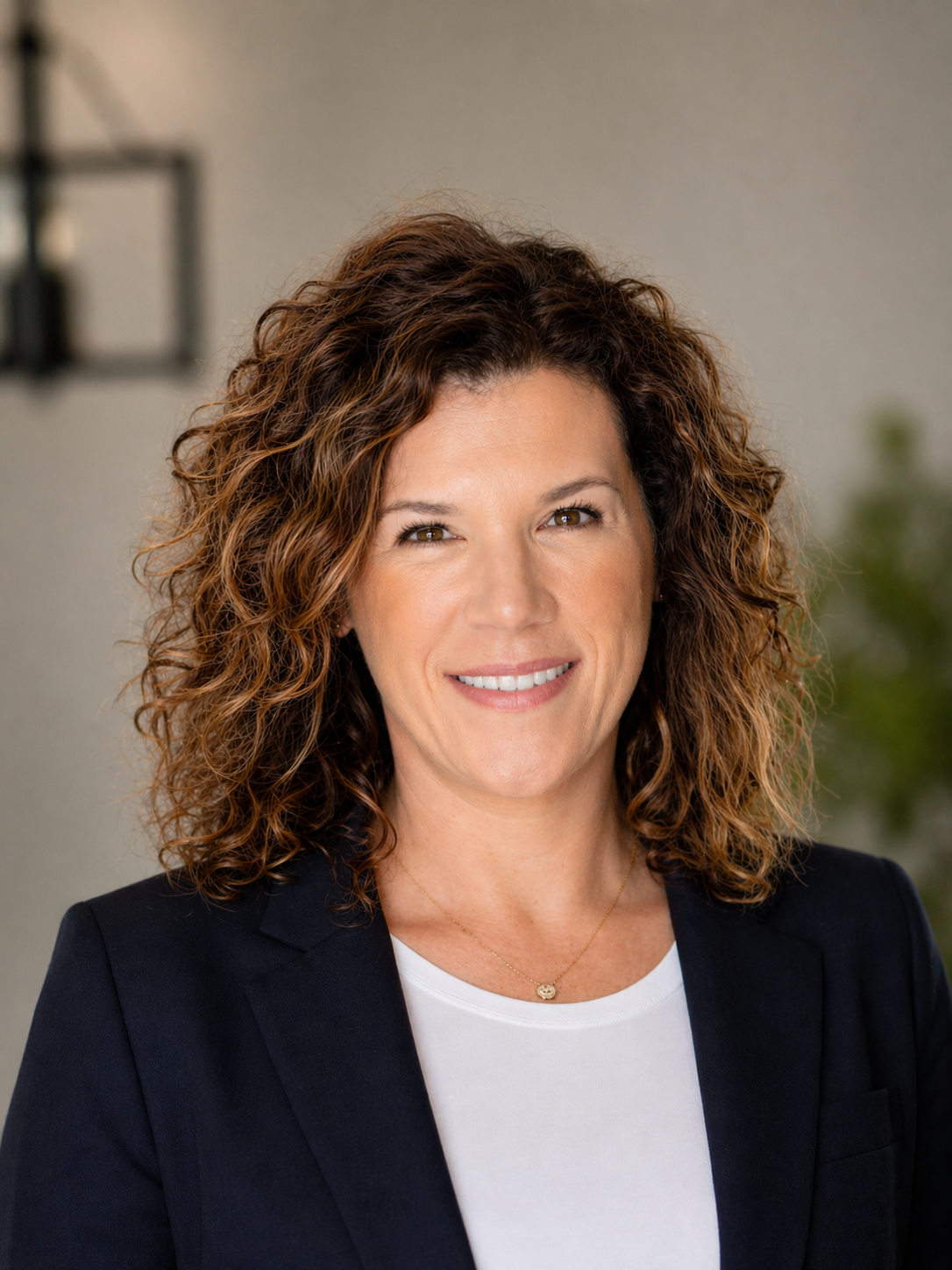 Professional headshot of a smiling woman with curly brown hair wearing a navy blazer and white shirt