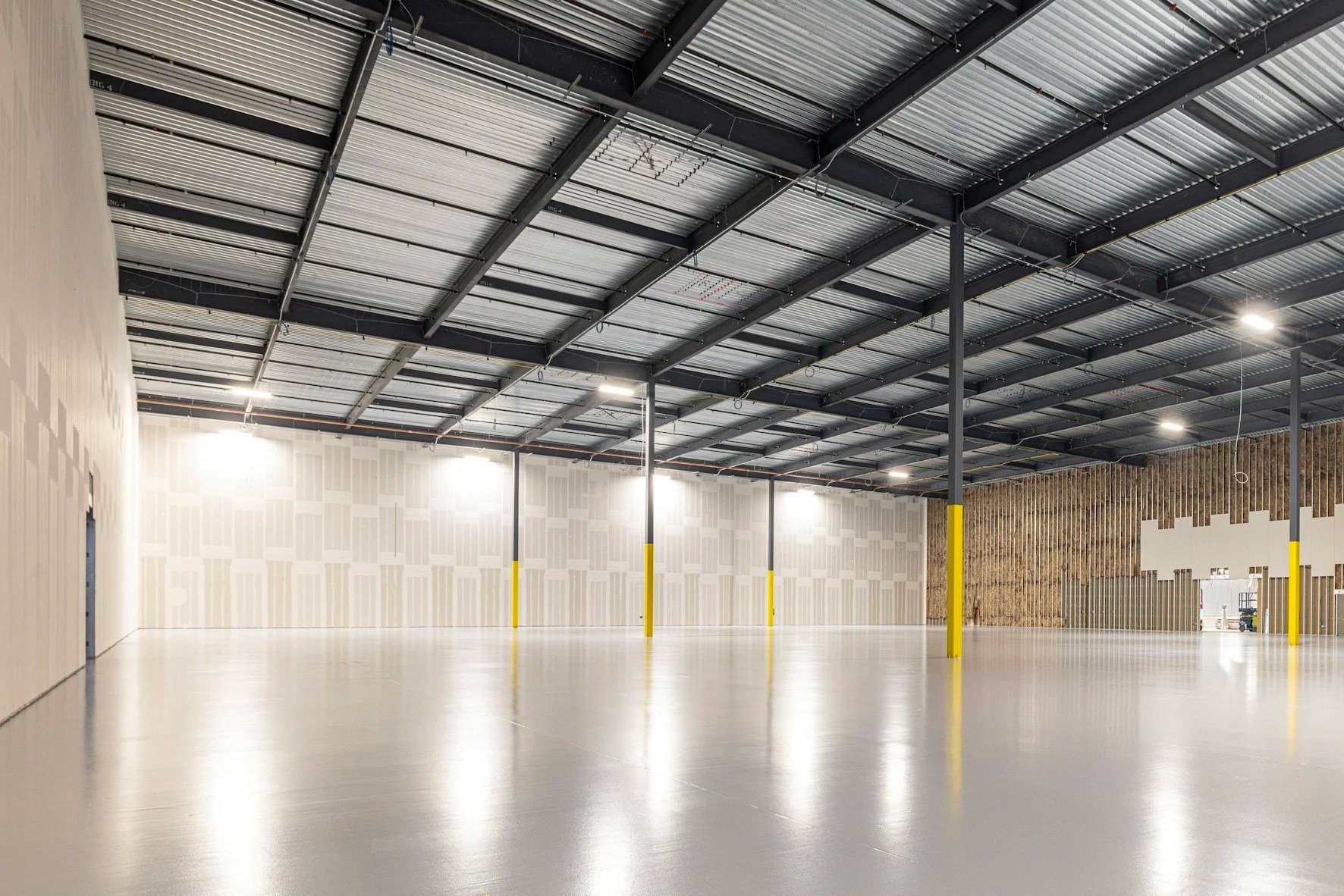 Empty modern warehouse interior with polished concrete floor, exposed steel roof beams, bright lighting, and yellow safety-column guards