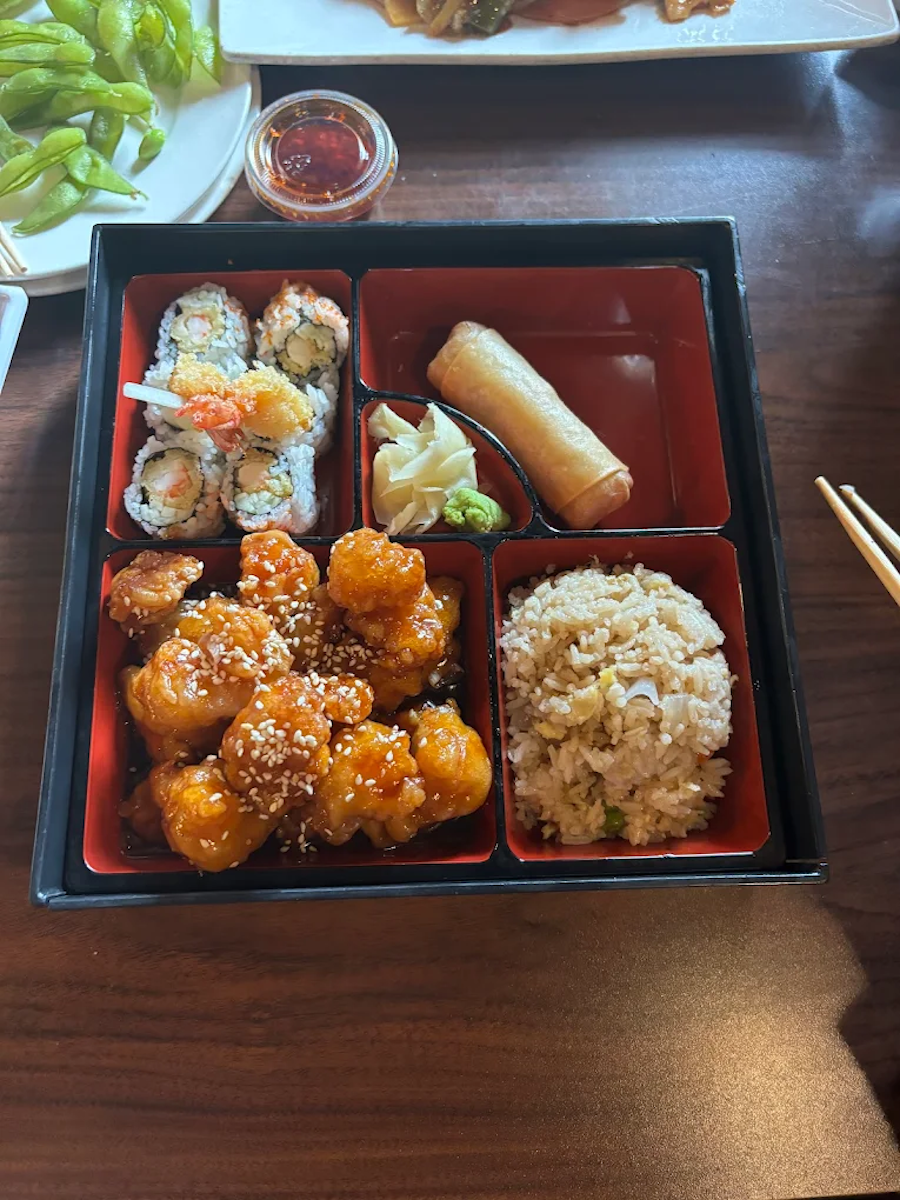 A bento box with sushi rolls, fried chicken pieces sprinkled with sesame seeds, a spring roll, a small portion of ginger and wasabi, and a portion of fried rice. A small container of soy sauce is also visible on the table.