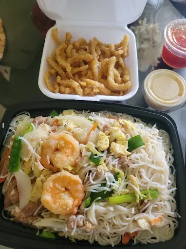 Takeout meal consisting of fried calamari in a white foam container, and a black tray of shrimp and vegetable lo mein with noodles and chopped green onions. Accompanying are two small containers of sauce and a cup of red beverage.