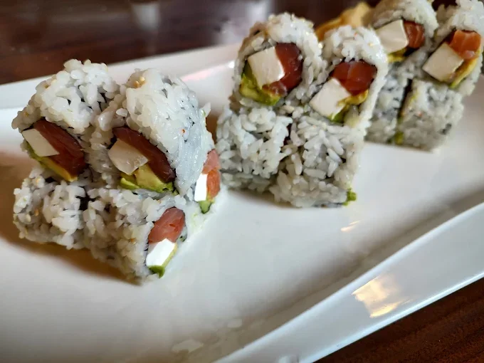 Close-up of sushi rolls with rice, salmon, cream cheese, avocado, and tuna on a white rectangular plate.