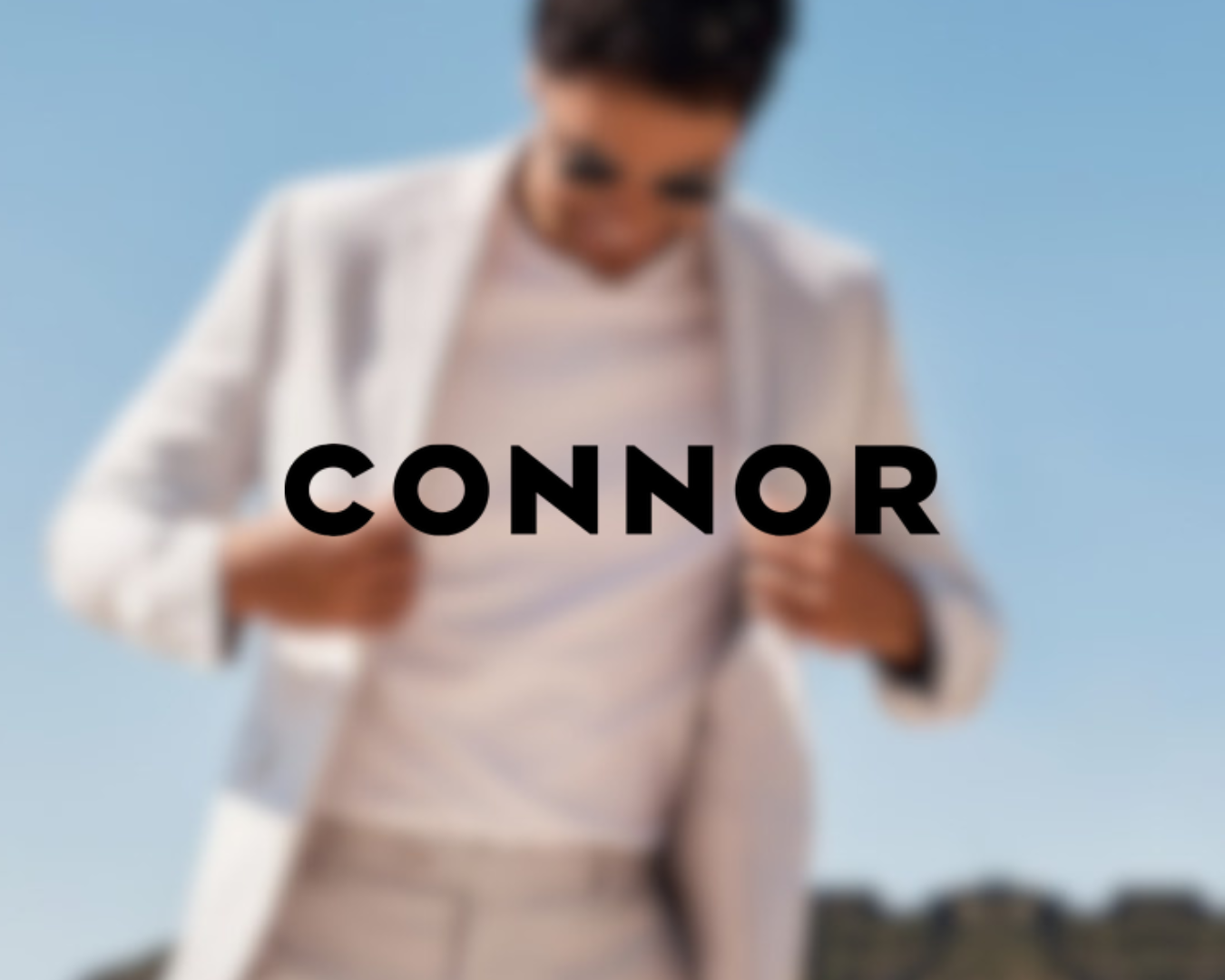 A person in a white blazer and beige pants looking down at a smartphone against a clear blue sky, with the name 'Connor' overlaid in bold black letters.