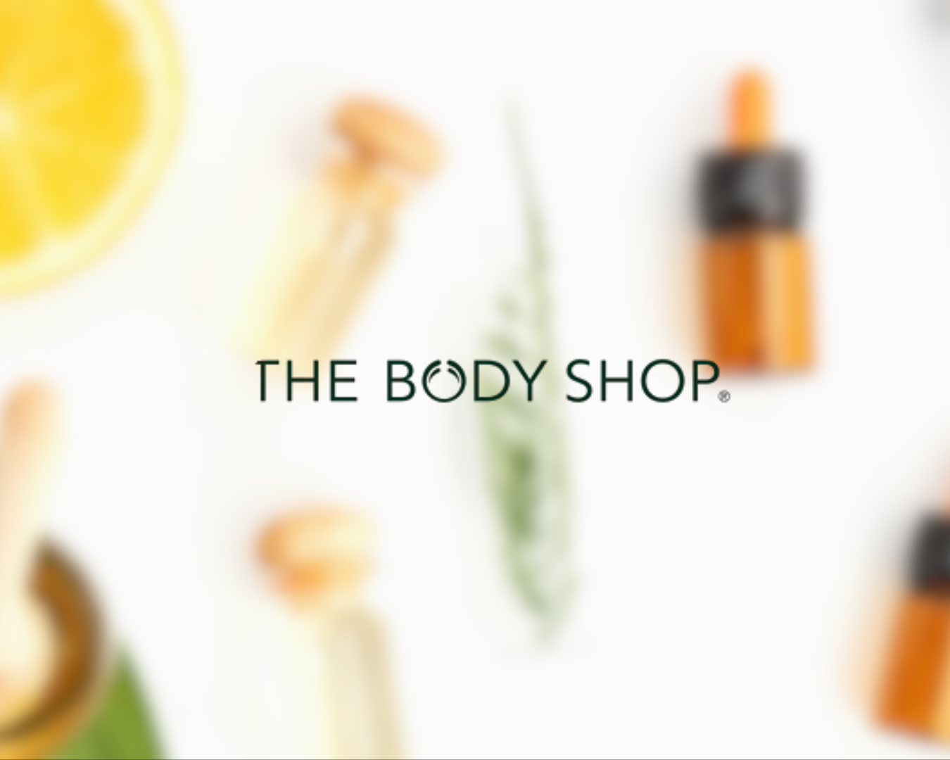 Blurred background with skincare products and a lemon slice, The Body Shop logo in the center.