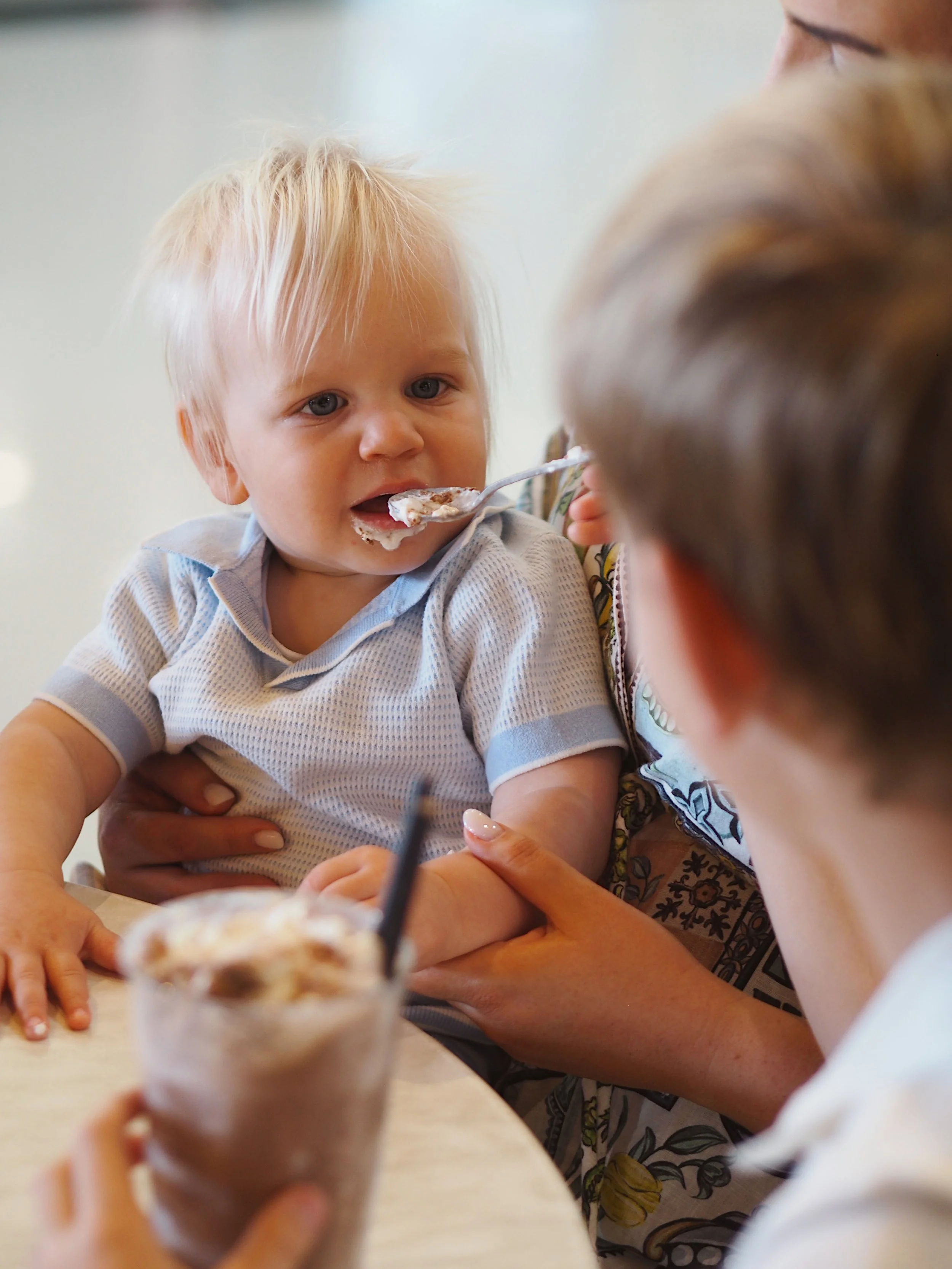 A young blond boy with blue eyes is being fed a spoon of ice cream by an adult. The boy is sitting at a table, and a blurred ice cream sundae with a cherry and straw is in the foreground. The adult's arm and shoulder are visible, and the background is plain and light-colored.
