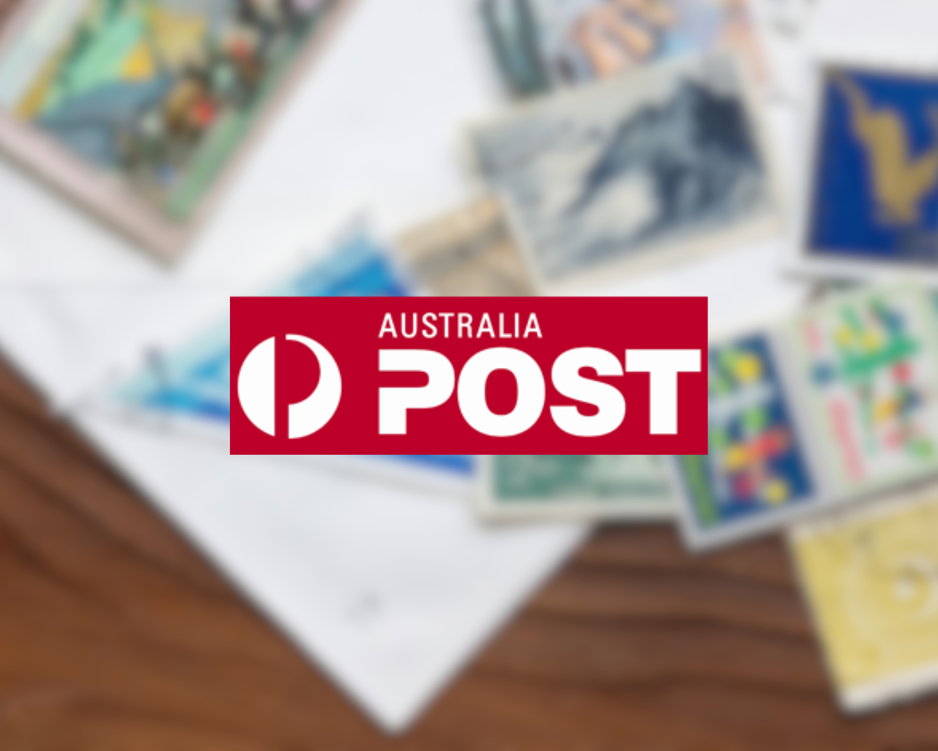 Blurred image of various Australian postage stamps and postcards on a wooden surface with a white sheet of paper underneath. A red Australia Post logo is overlaid in the center.