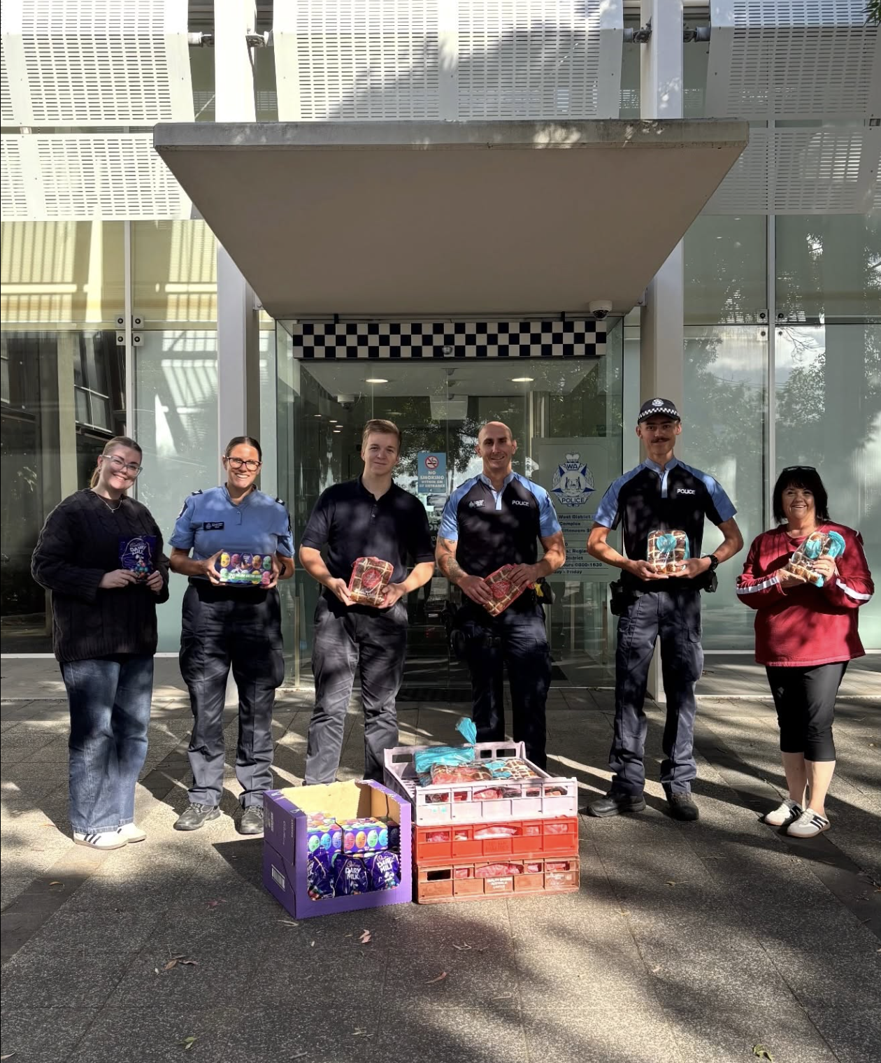 Group of six people, including police officers and civilians, standing in front of a building entrance, holding wrapped gifts and standing behind crates filled with snacks and candies.