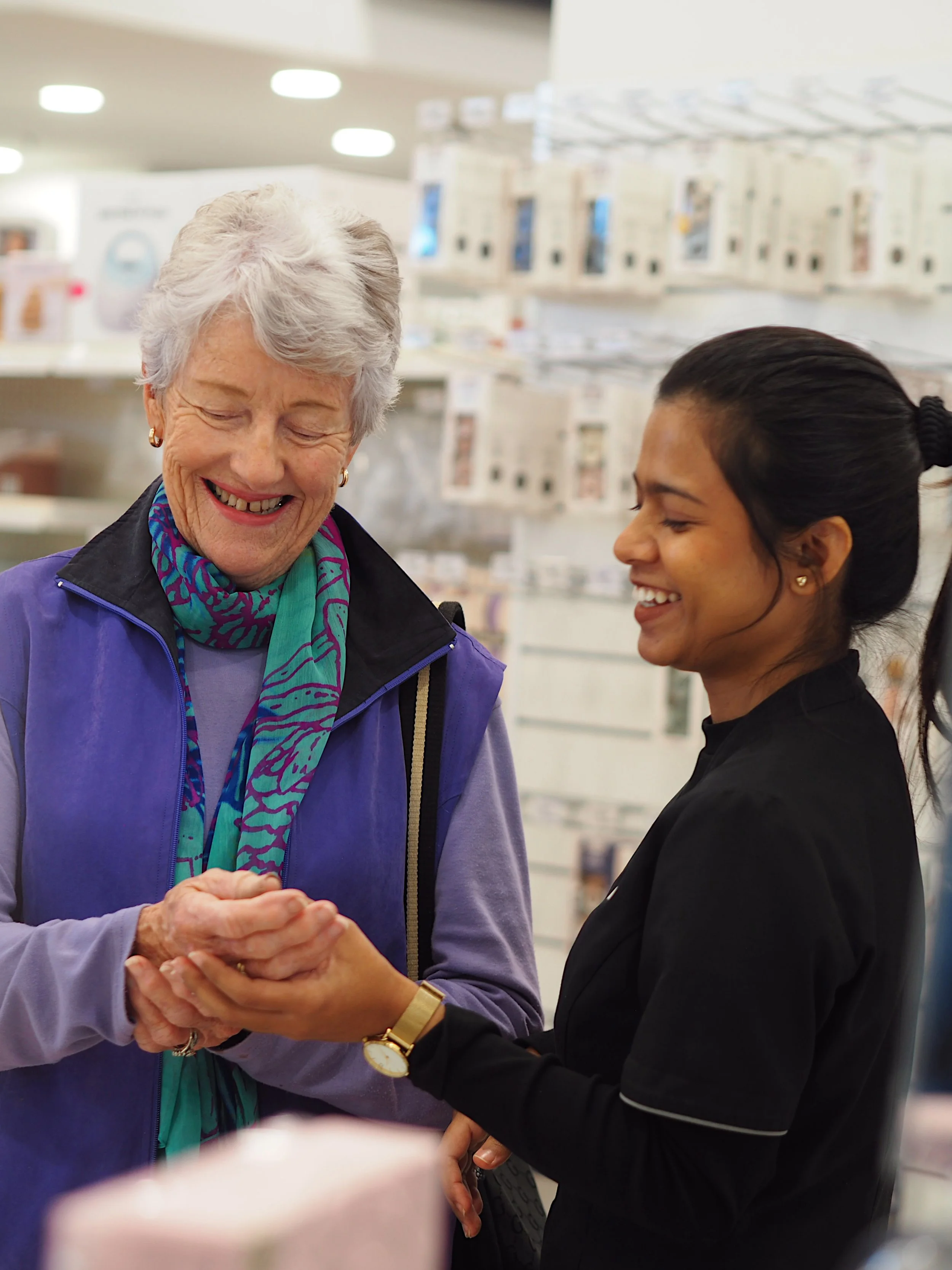 An elderly woman with white hair and a smiling face, wearing a purple jacket and a colorful scarf, holding hands and smiling with a young woman with dark hair tied back in a ponytail, dressed in a black top, in a retail store with shelves of products in the background.