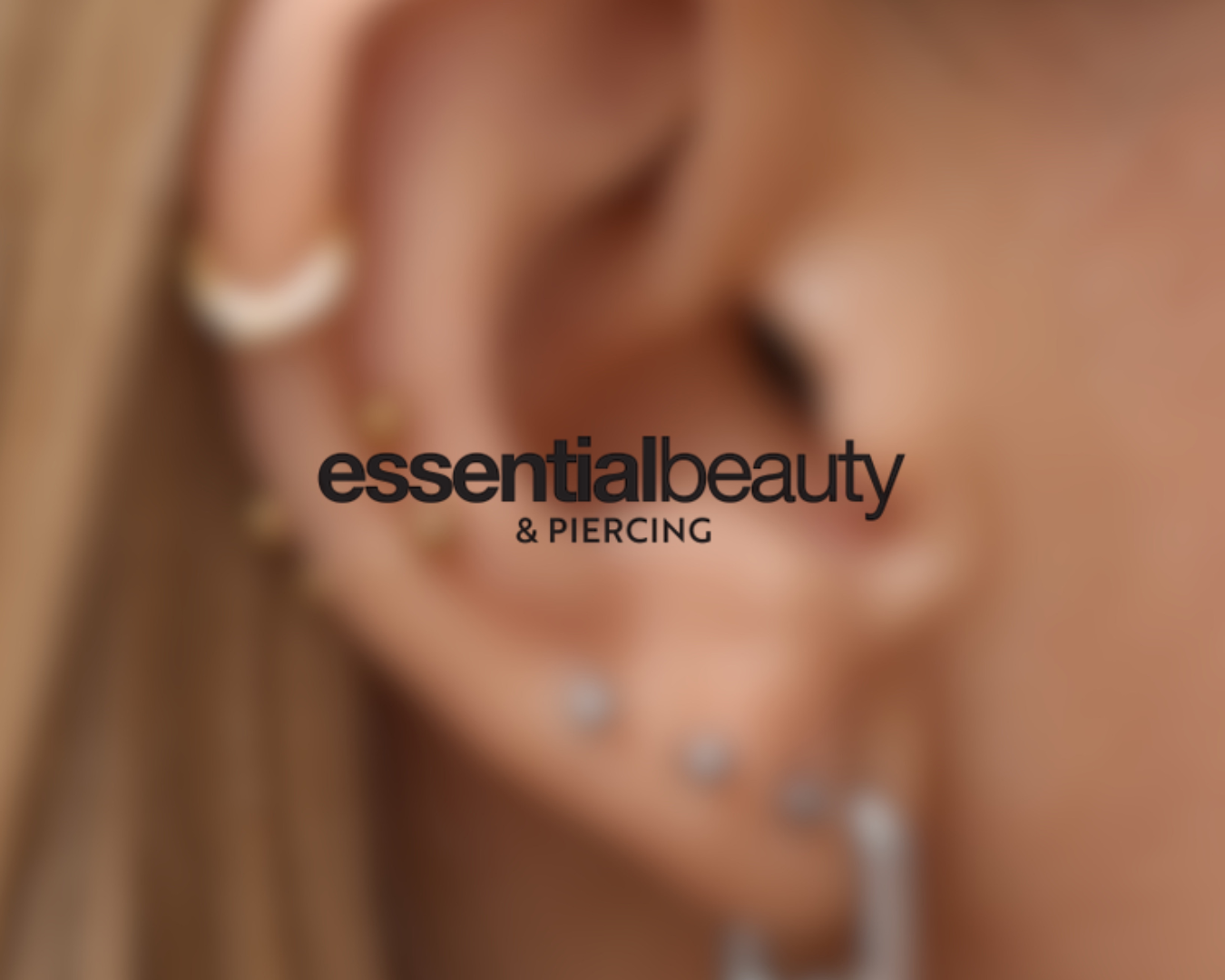 Close-up of a woman with multiple facial piercings, wearing earrings, with the text 'essential beauty & piercing' overlaid.