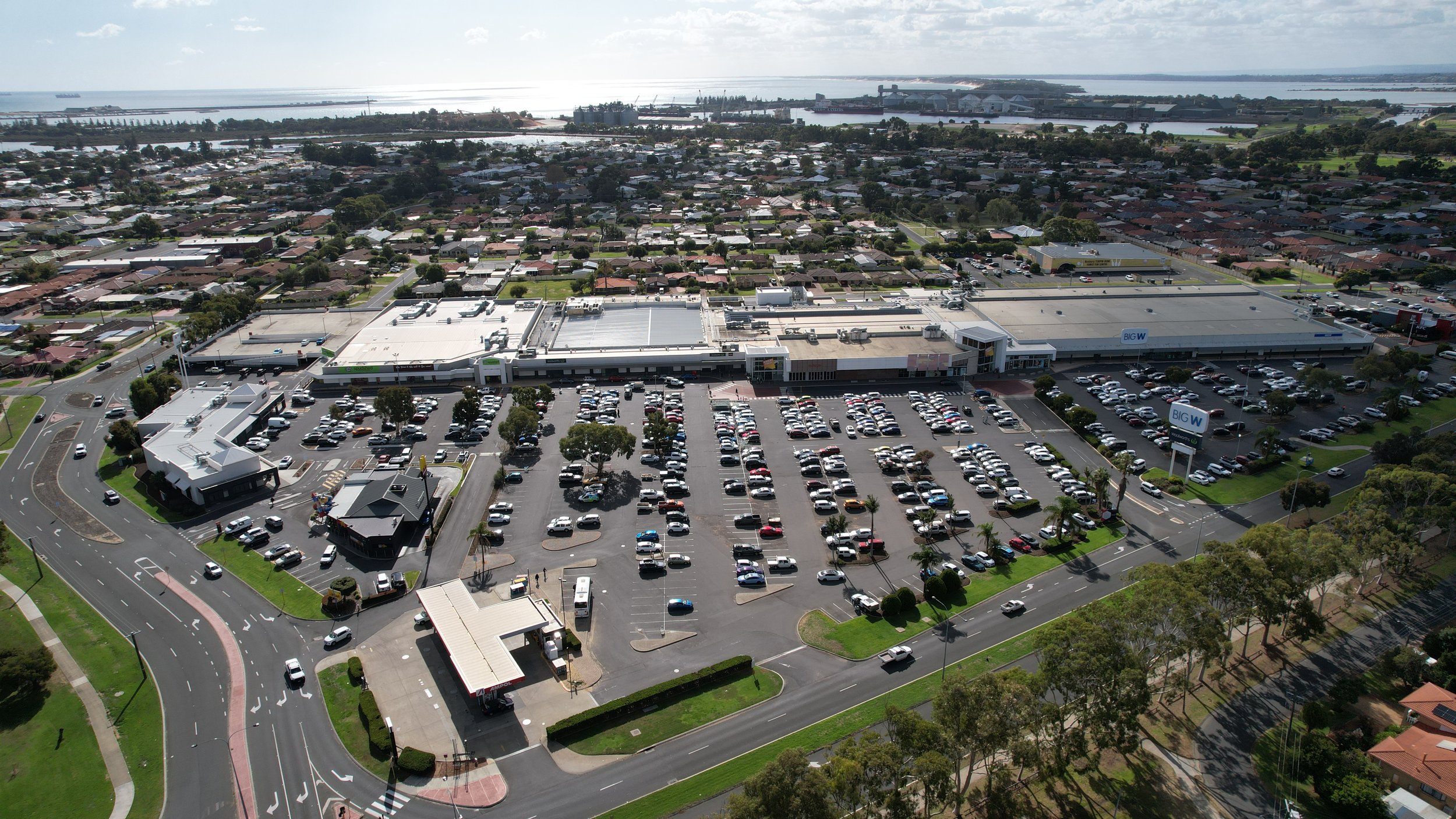 Aerial view of a shopping center with parking lot filled with cars, surrounding roads, residential houses, and a body of water in the background.