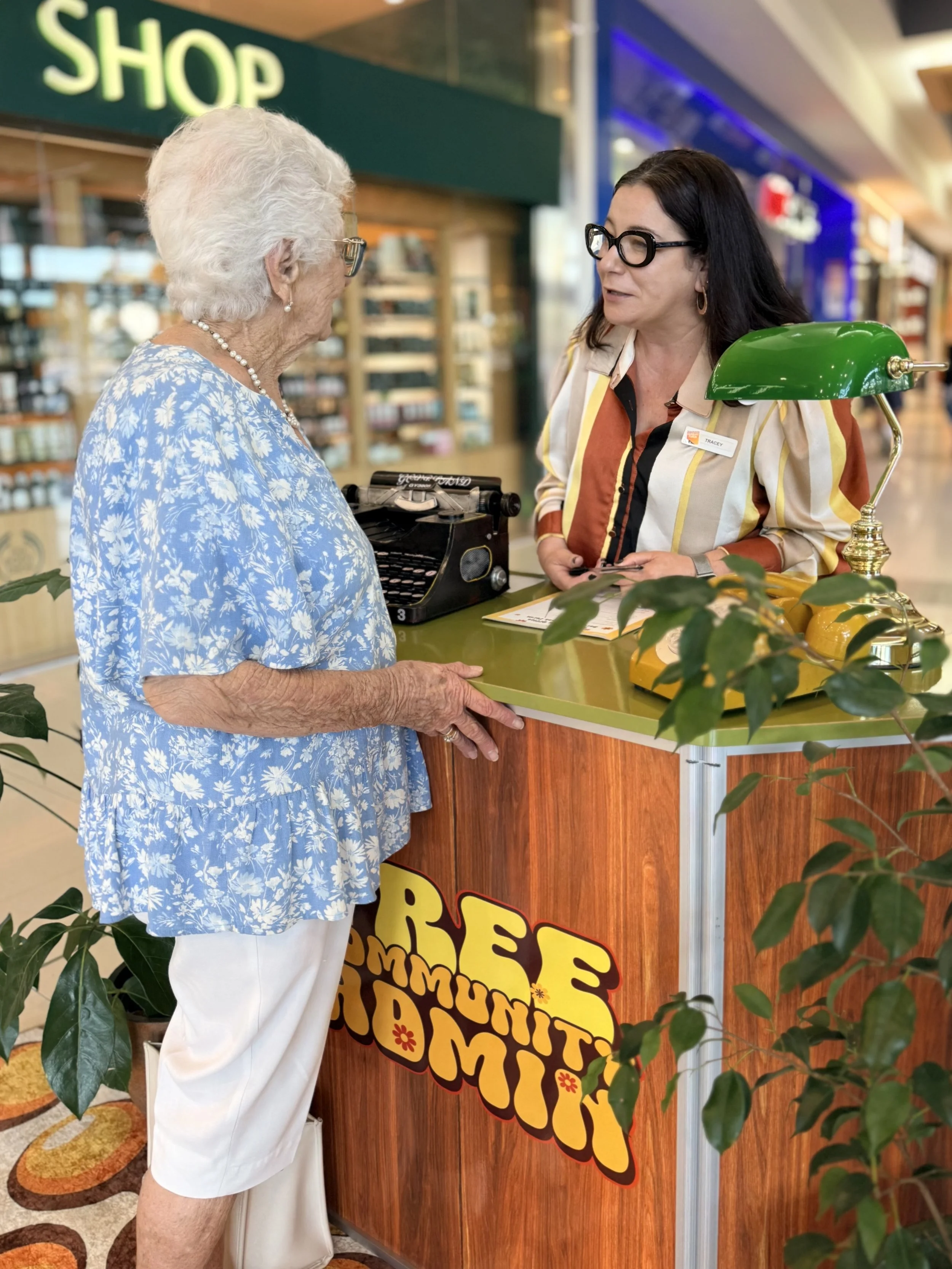 An elderly woman with gray hair wearing a blue floral dress is talking to a cashier behind a colorful sign that reads 'SUMMER SPECIAL'. The cashier is a woman with long dark hair and glasses, wearing a multicolored blouse, at a retail counter in a shopping mall.