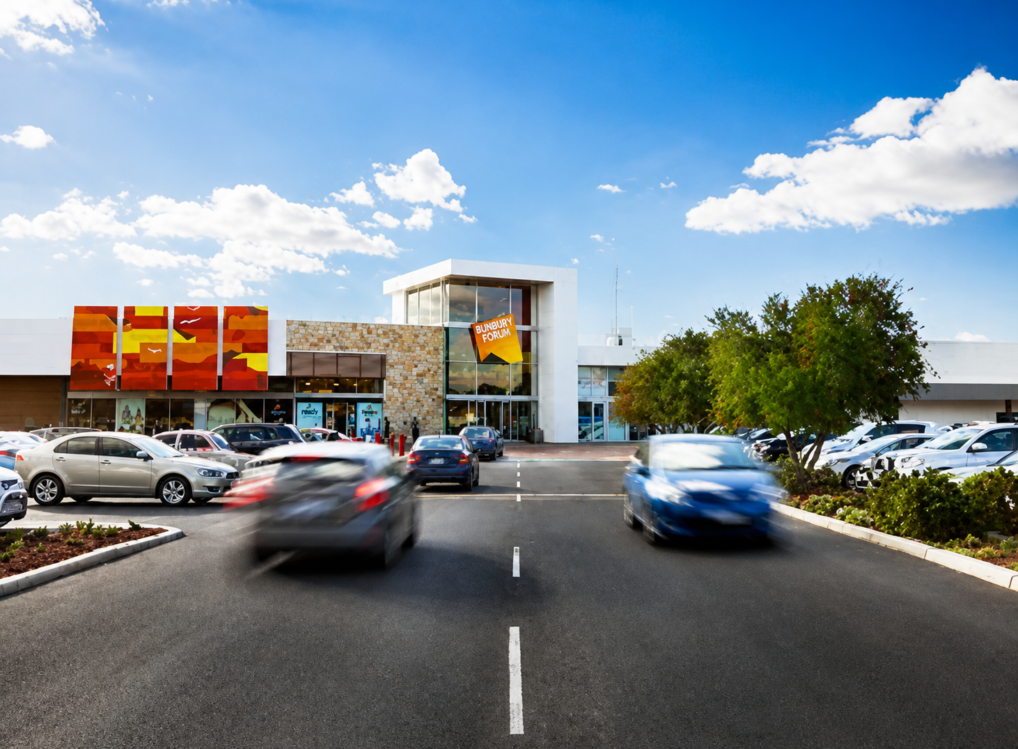 Parking lot in front of Bunbury Forum shopping mall with cars and trees on a sunny day.