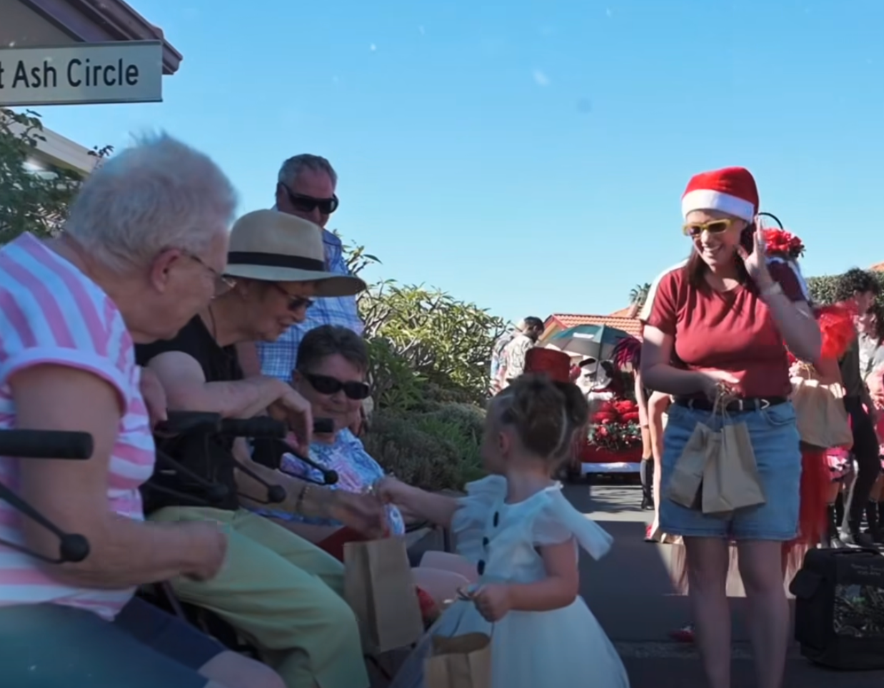 A group of elderly women sitting on benches and children interacting with them at a festive outdoor event. One woman wears a Santa hat, and there are holiday decorations in the background.