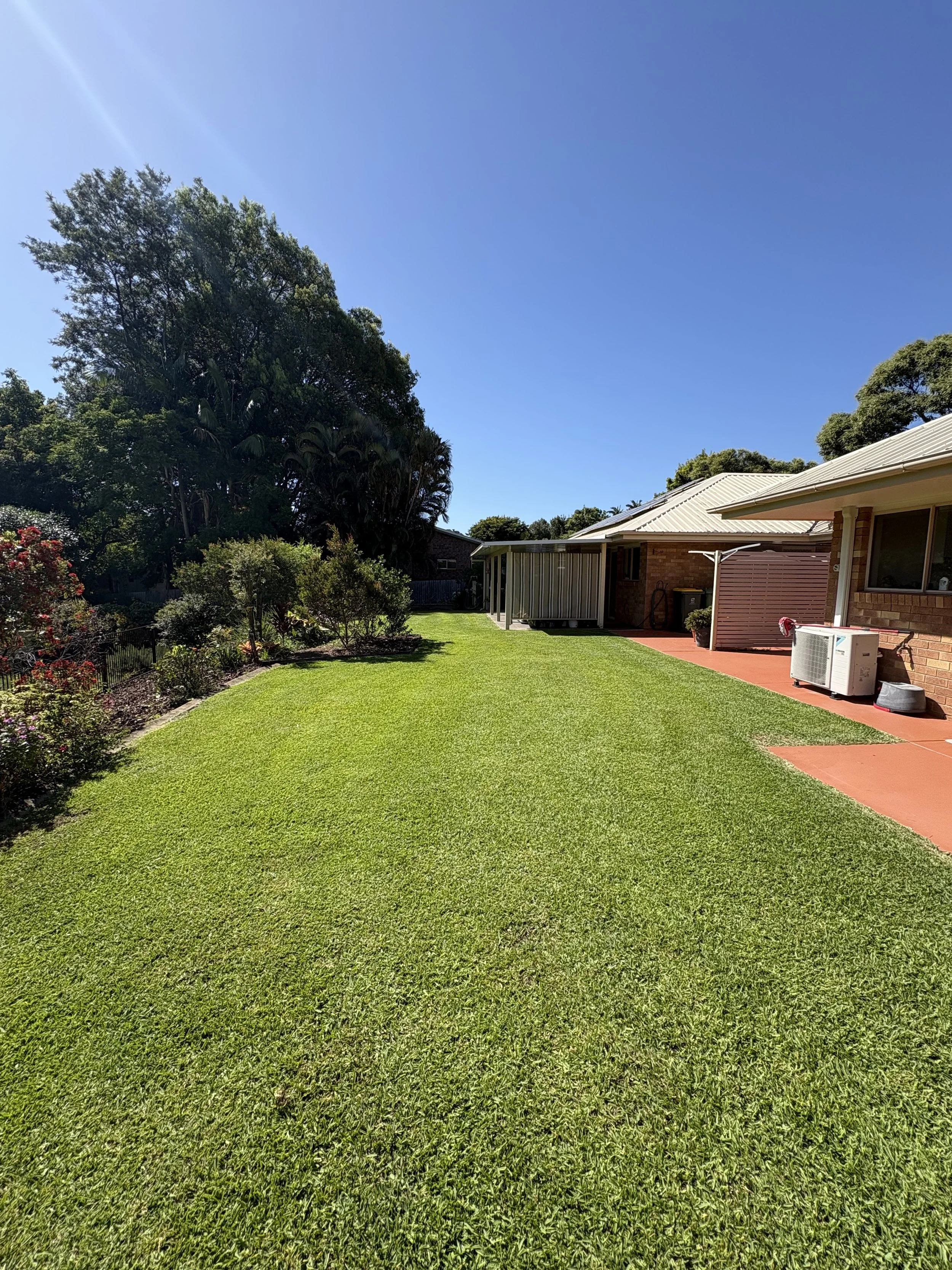 Well-maintained backyard with lush green grass, trees, and shrubs, along with a brick house featuring a metal roof and a concrete patio area.