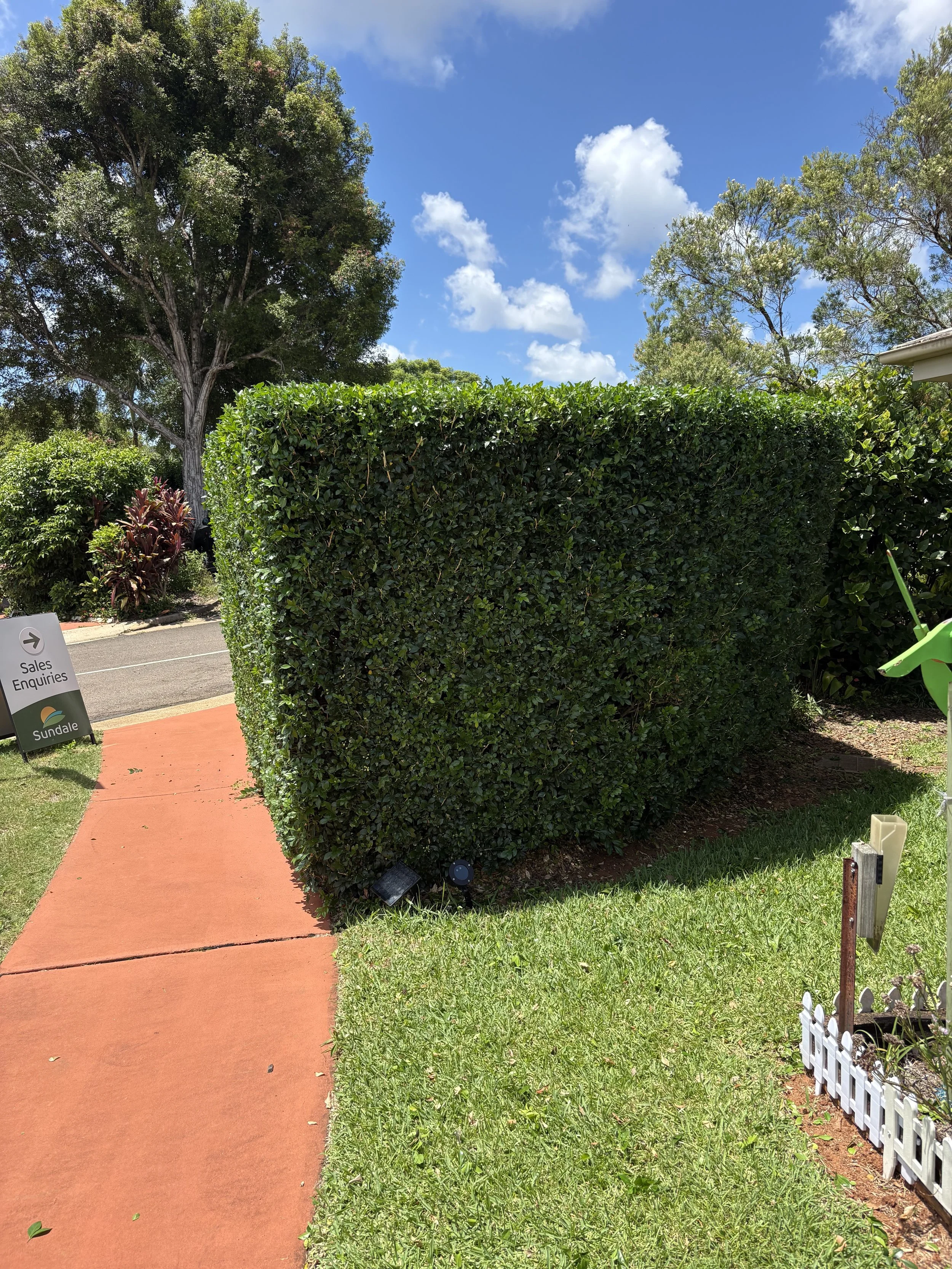 A sidewalk leads past a large, neatly trimmed green hedge in a residential area with a lawn, trees, and a sign that reads 'Sales Enquiries.' The sky is blue with white clouds.