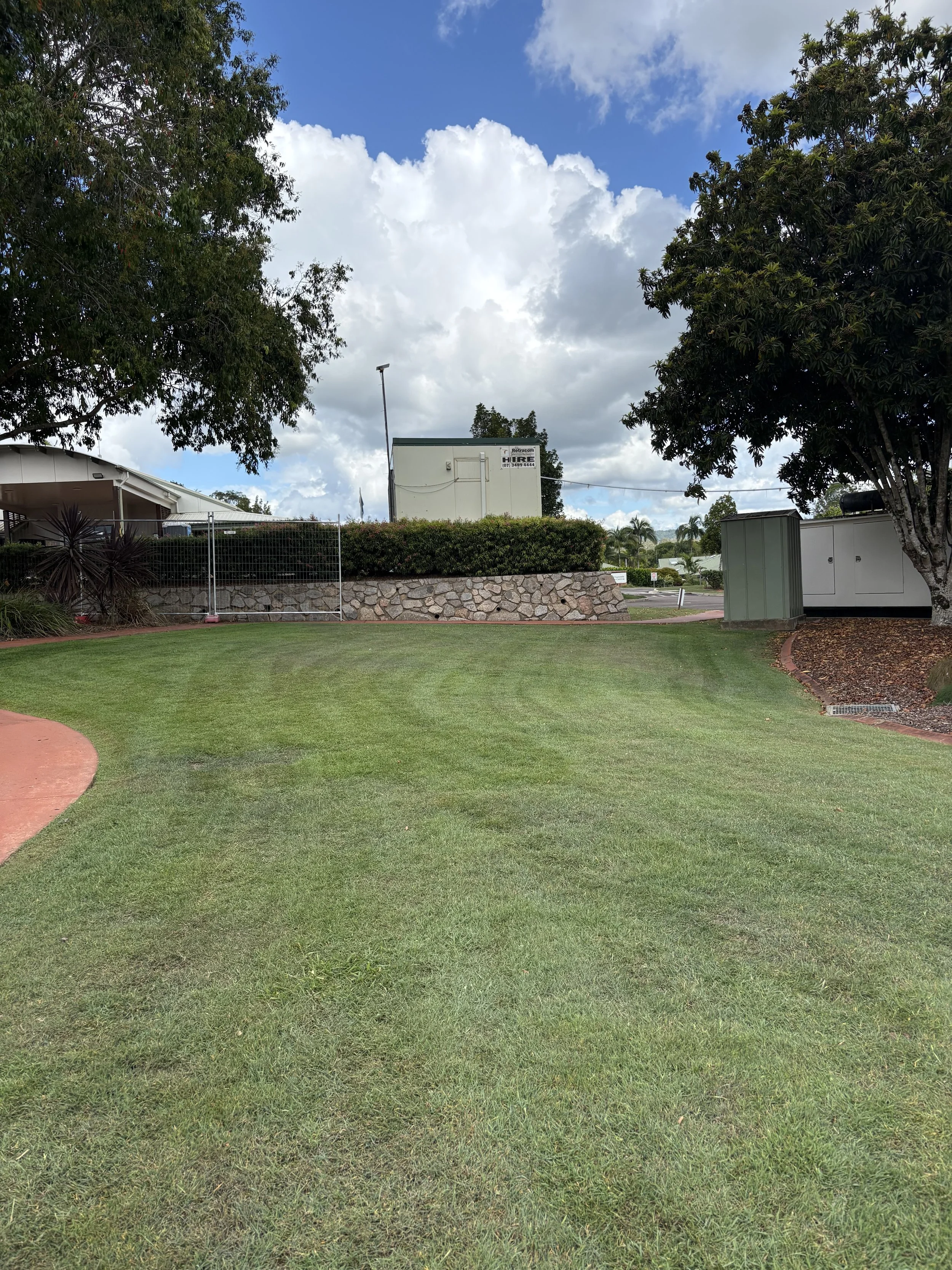 Well-manicured grass lawn with a curved red pathway on the left side, trees on either side, a stone wall behind the lawn, and a utility building in the background under a partly cloudy sky.