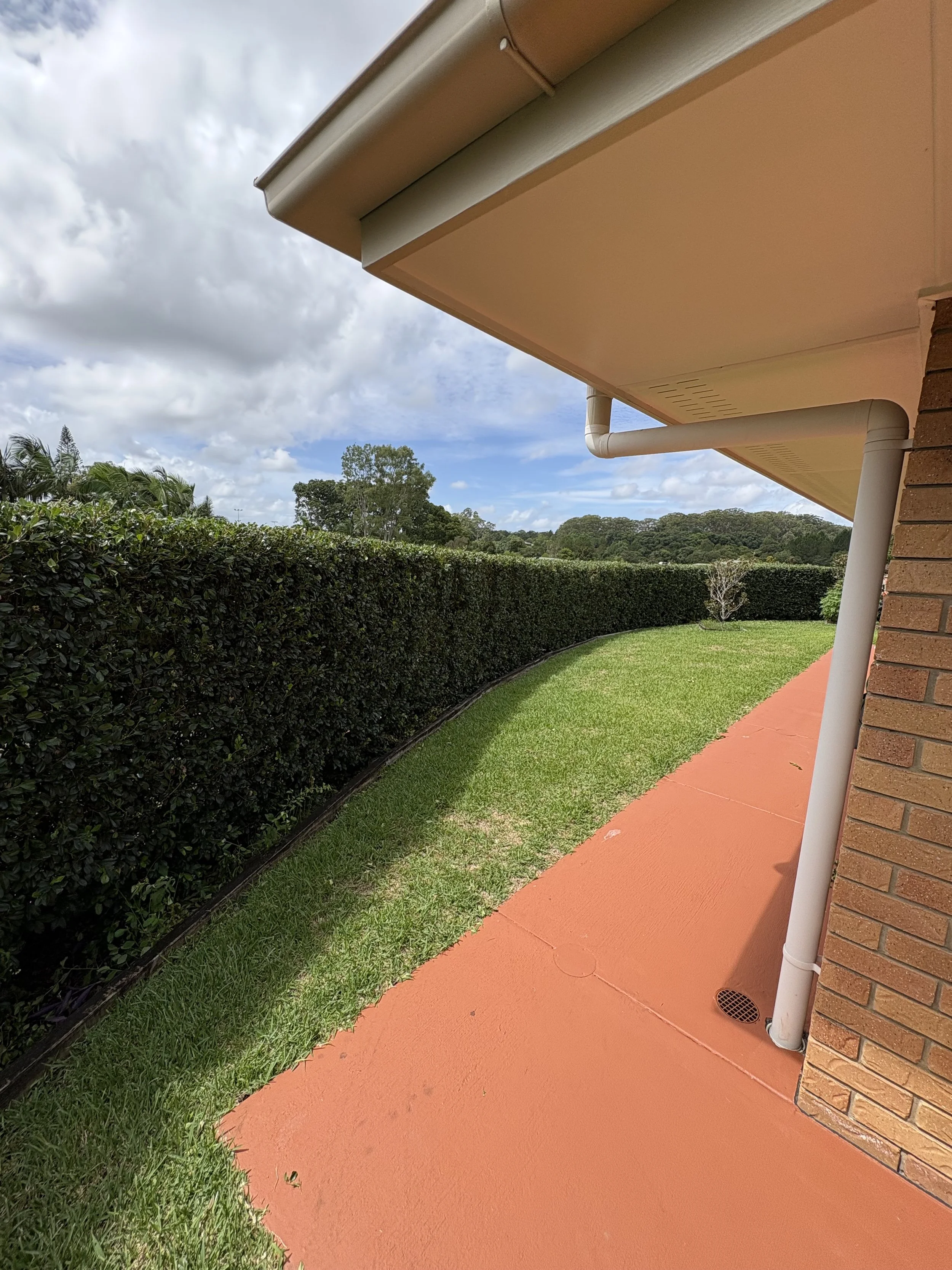Side view of a suburban house with a brick wall, rain gutter, white downspout, clean orange concrete walkway, lush green shrubs, and a partly cloudy sky.