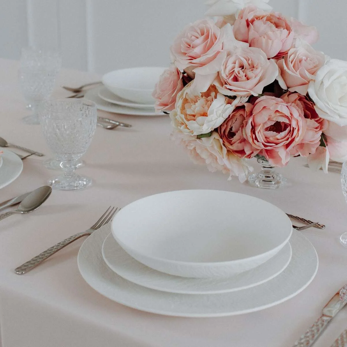 Elegant table setting with white plates, silverware, crystal glasses, and a floral centerpiece of pink and white roses.