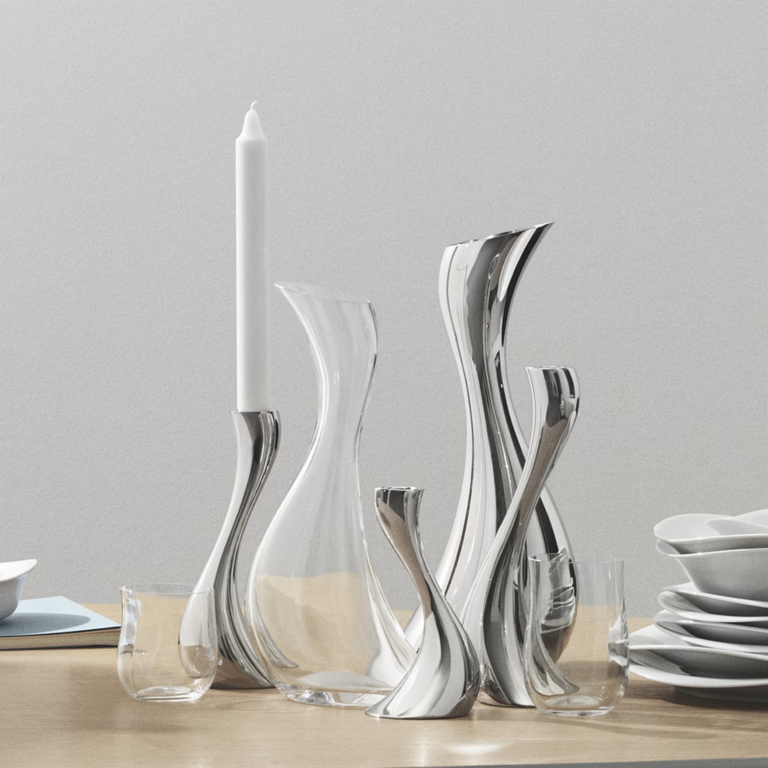 Various empty glassware including drinking glasses, a decanter, a wine glass, and a carafe arranged on a wooden surface with a grey background.