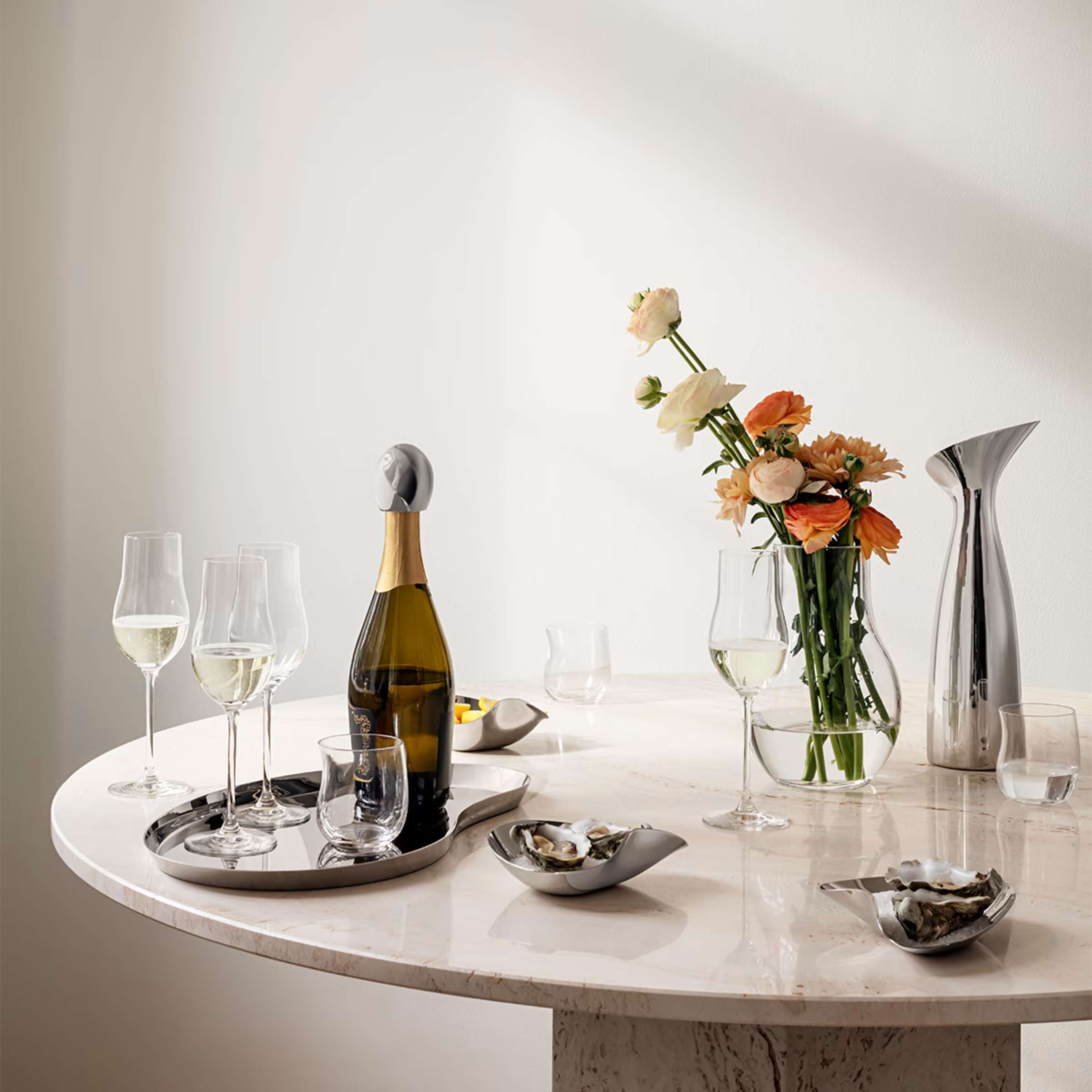 A white marble table with a silver tray holding wine glasses, a champagne bottle, and various bowls with oysters and shellfish. There is a glass vase with a bouquet of pink and orange flowers, and a set of silver candlesticks with black candles in the background.