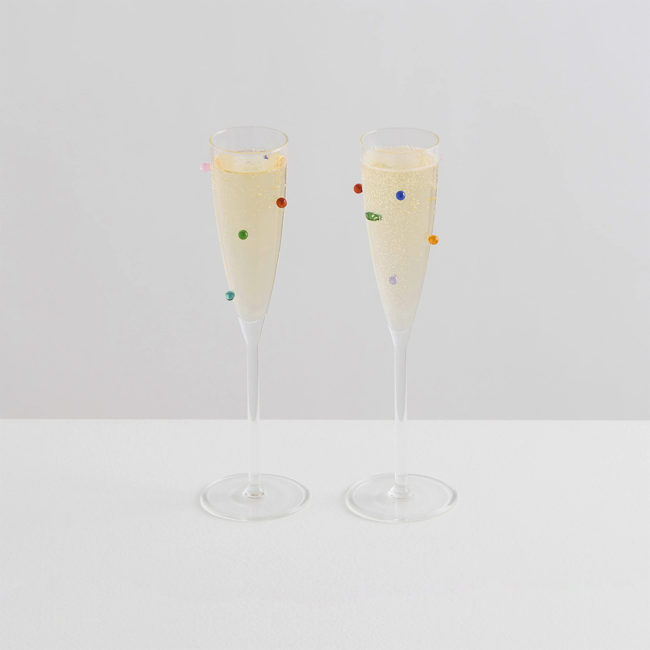 Two tall champagne flutes filled with sparkling wine, decorated with small colorful beads hanging on the outside, set against a plain white background.
