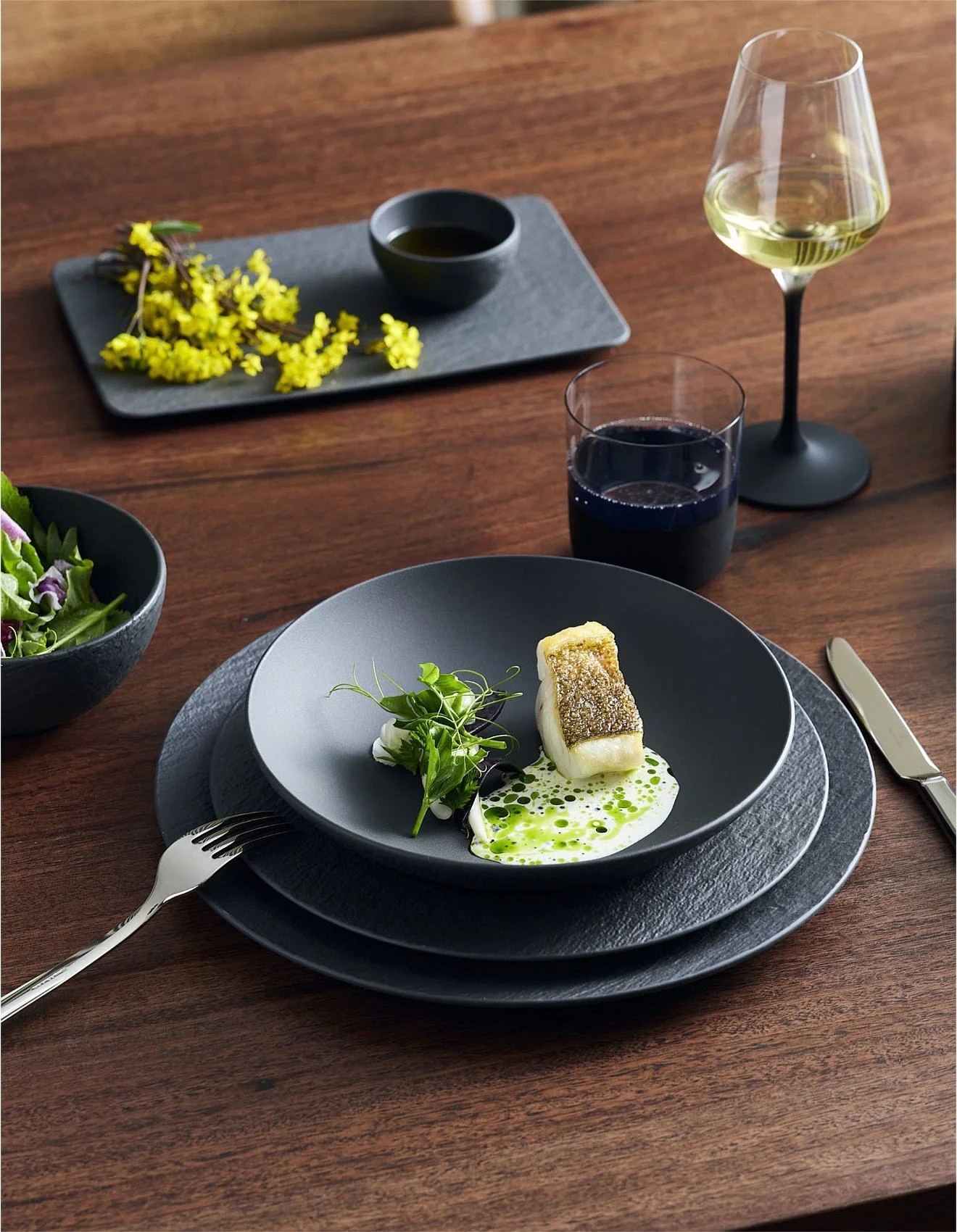 A plated gourmet dish featuring a piece of fish with crispy skin, garnished with greens and a green sauce, served on a black plate with a fork and knife. Behind the main dish are glasses of white and red wine and a small bowl of salad. To the side, there is a black bowl of greens and a rectangular tray with a small black bowl of dipping sauce and yellow flowers, all on a wooden table.