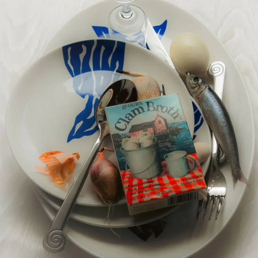 A white plate with a blue design holds a box of clam broth, a spoon, an onion, and a fish, with a seashell on the plate and a drinking glass with water behind the plate.