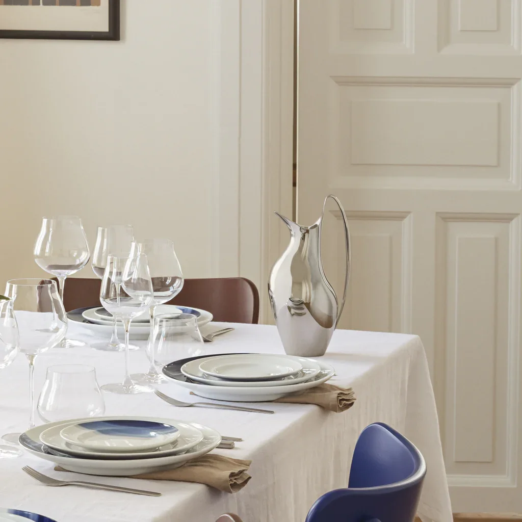 A dining table set for a meal with white plates, silverware, wine glasses, a silver pitcher, and a beige napkin on a white tablecloth. Part of a blue chair and a brown chair are visible, with a white door in the background.