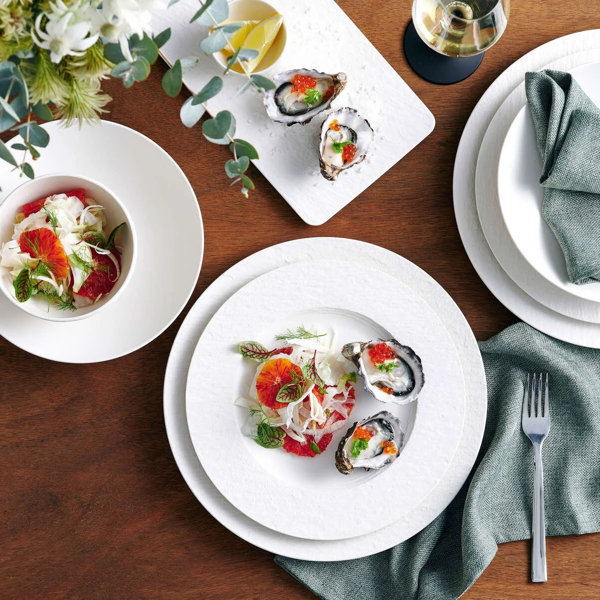 A table setting with oysters topped with red caviar and herbs, a bowl of salad with grapefruit segments and greens, and a glass of white wine, all arranged on white plates and a wooden table surface.