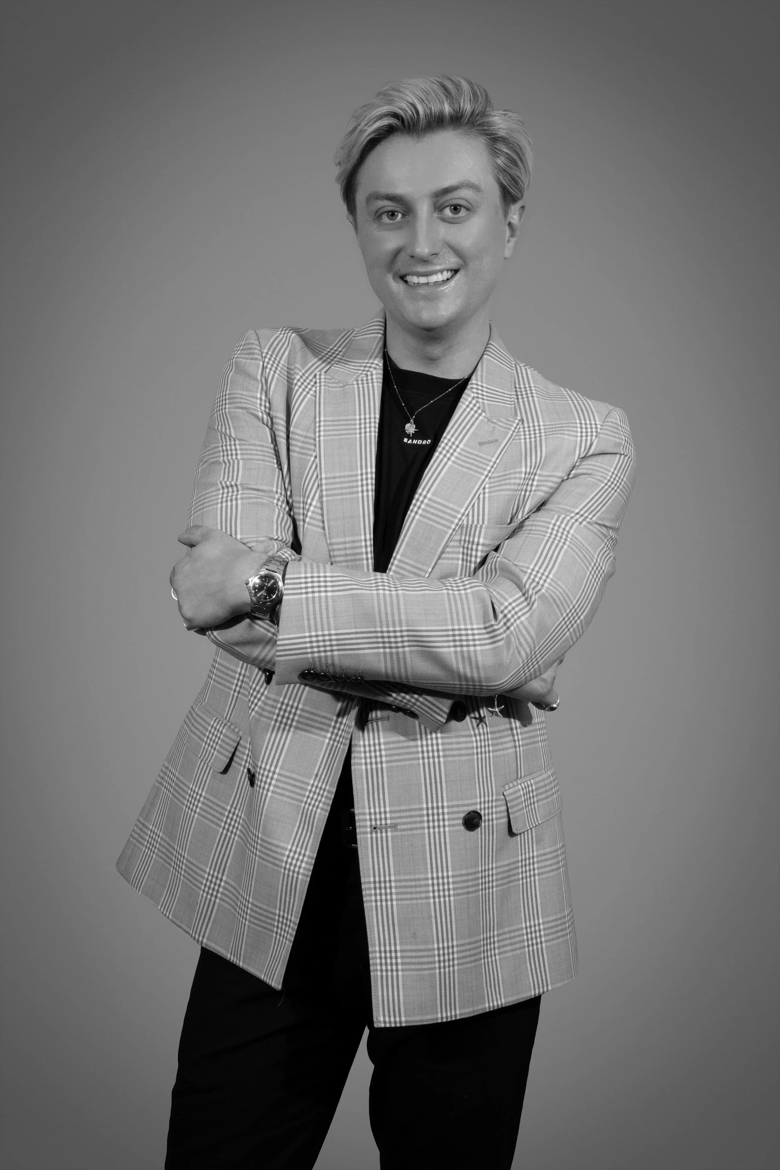 Black and white portrait of a smiling young man with styled blonde hair, wearing a plaid blazer, a black shirt with the word 'SANDRO' on a necklace, a wristwatch, and black pants, crossing his arms against a plain background.