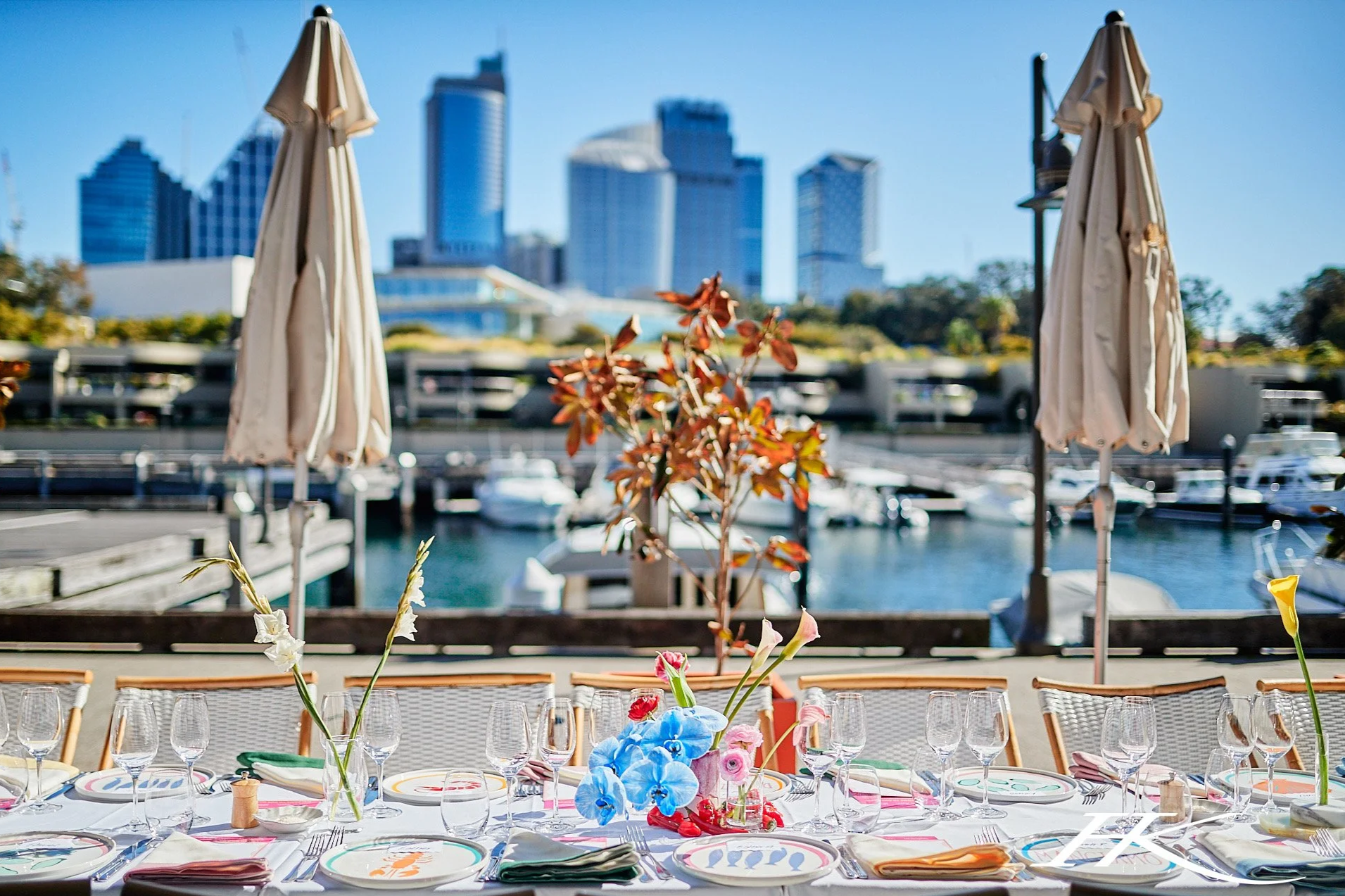 Table set for a celebration with colorful flowers and plates, overlooking a marina with boats and a city skyline with tall buildings in the background.