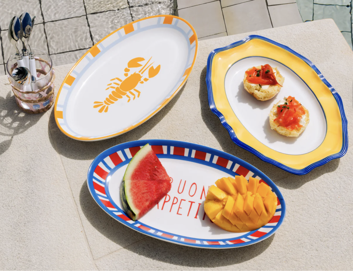 Colorful outdoor table with three decorative plates containing watermelon, mango, and two pieces of toast topped with salmon and herbs. There is a lobster-themed plate and a glass containing utensils.