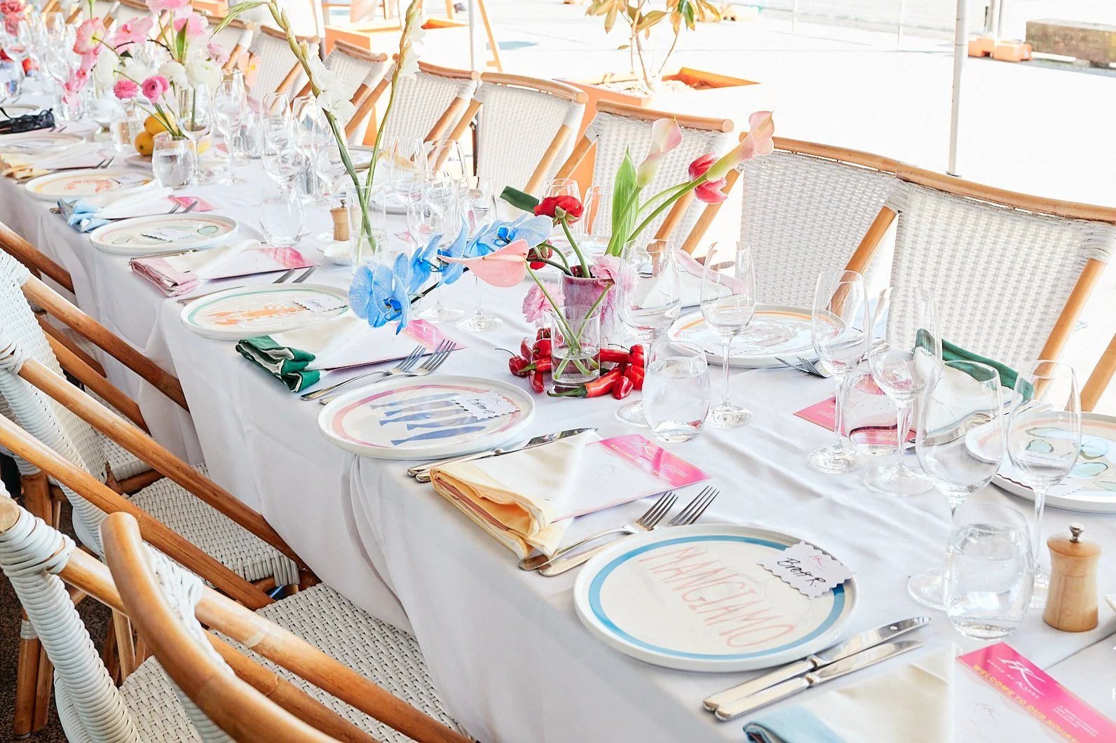 A long banquet table set for a celebration, decorated with floral centerpieces, colorful plates, napkins, and glassware, with chairs on both sides.