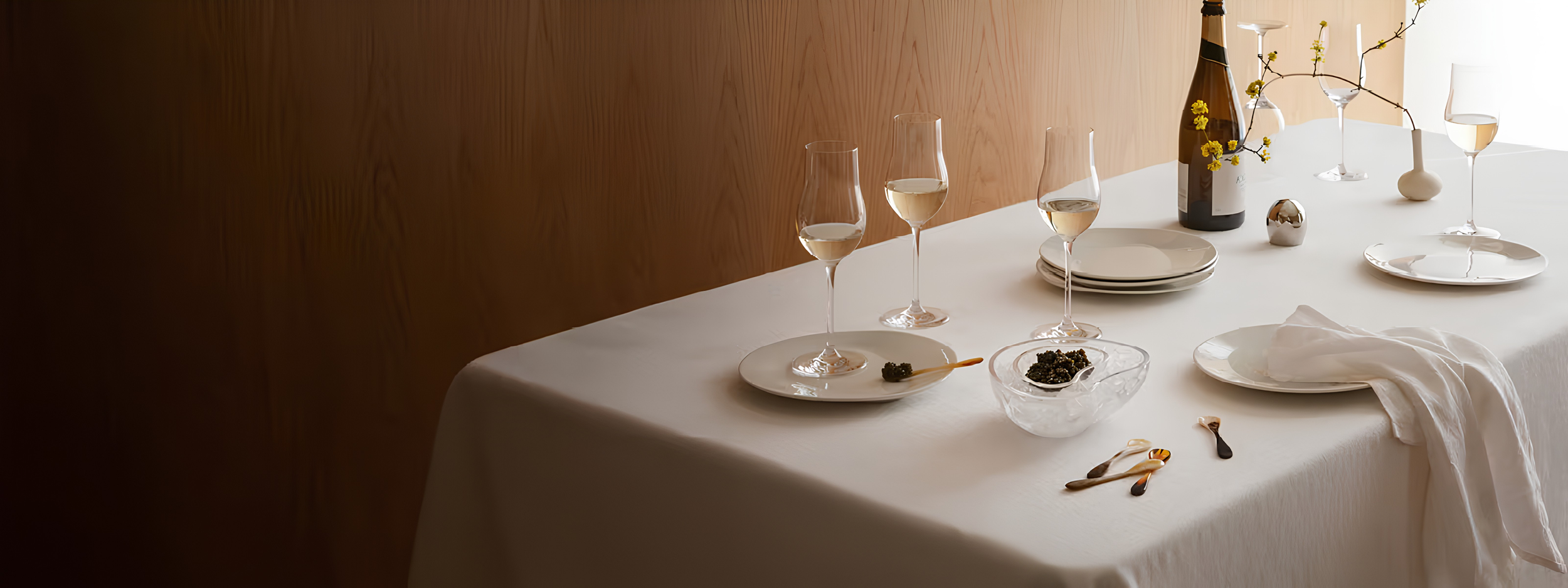 A dining table set with champagne glasses, a bottle of champagne, plates, a bowl of caviar, a vase with yellow flowers, and silverware on a white tablecloth.