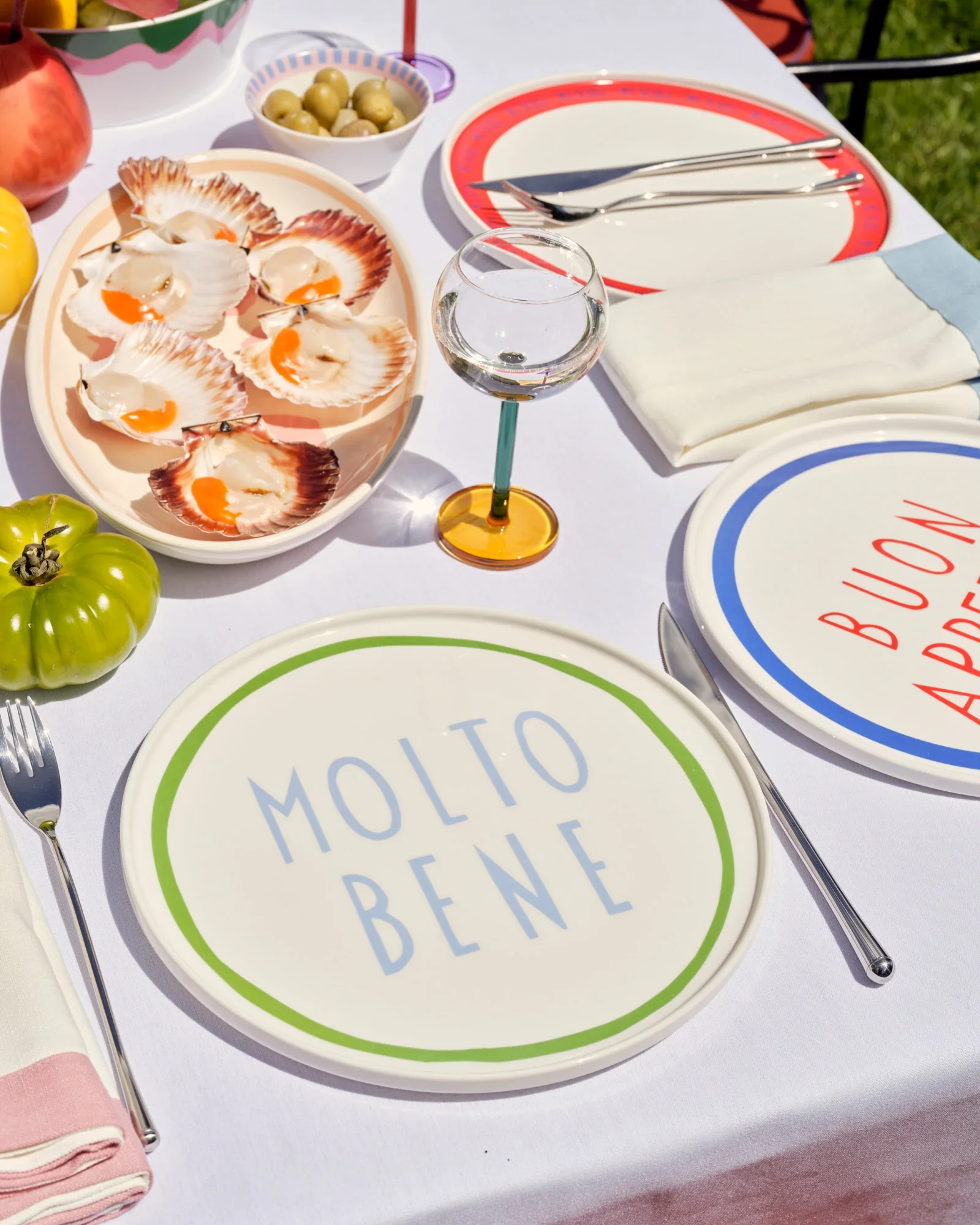A table set for a meal with a white tablecloth, featuring a plate of baked oysters, a small bowl of green olives, a glass of water, cutlery, a striped paper plate, two folded napkins, and some fresh vegetables including a green tomato and a yellow peach.