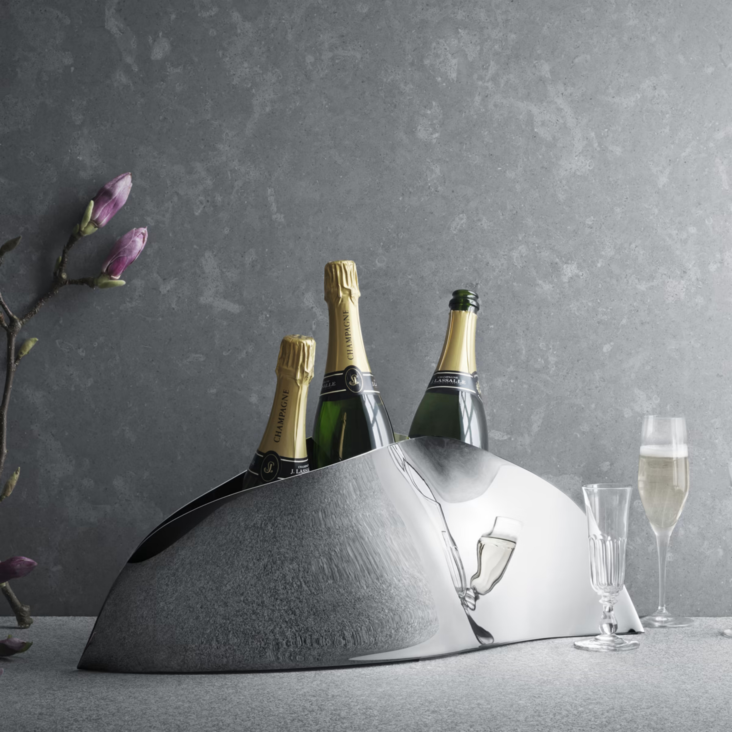 A silver ice bucket with champagne bottles, surrounded by empty champagne glasses, with a branch of pink magnolia flowers on the left side, set against a dark textured background.