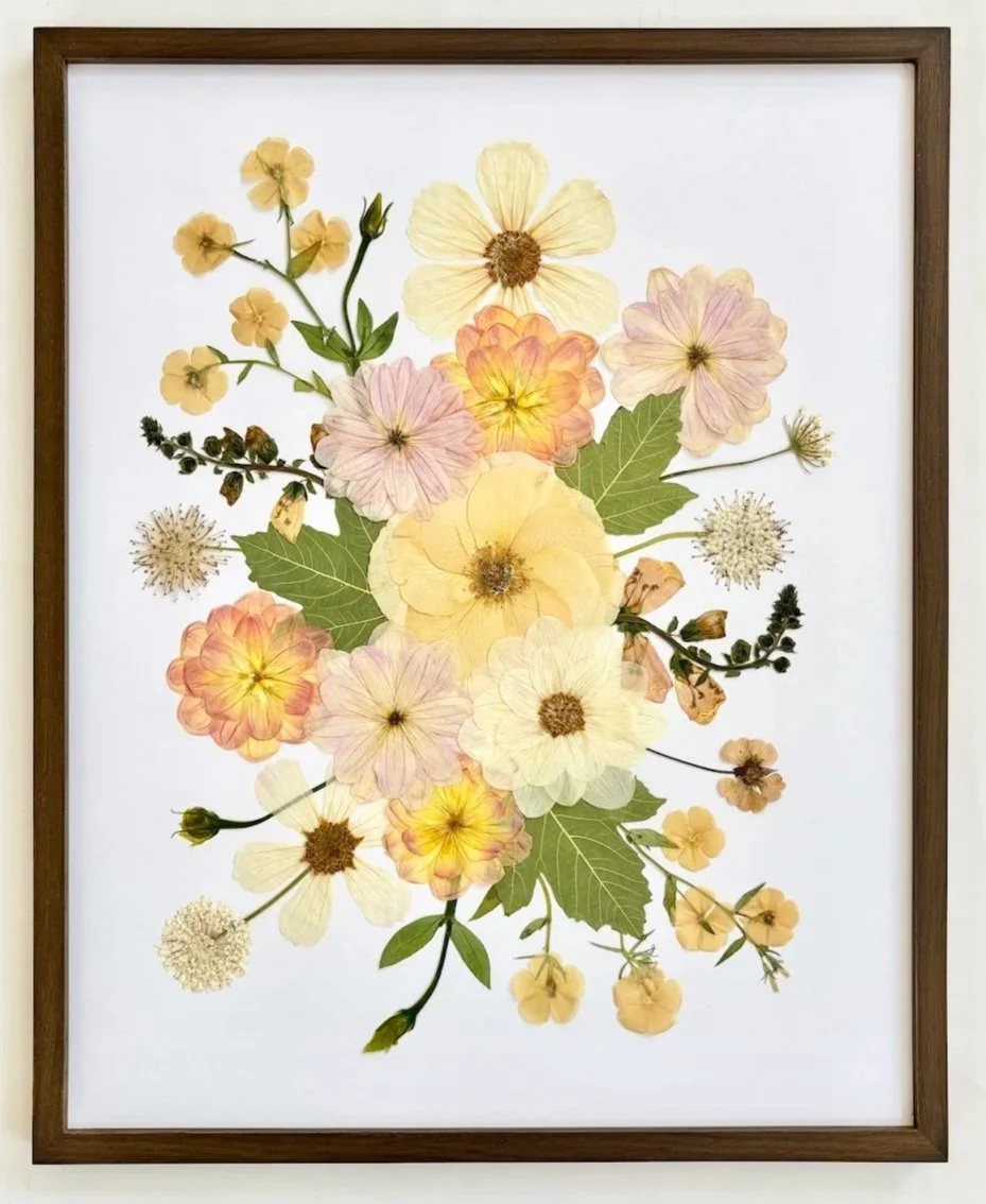 Framed botanical print featuring pressed flowers and leaves in soft pastel tones on a white background.