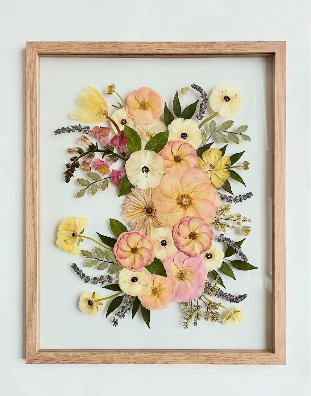 Framed wall art with pressed pastel-colored flowers and green leaves on a white background.