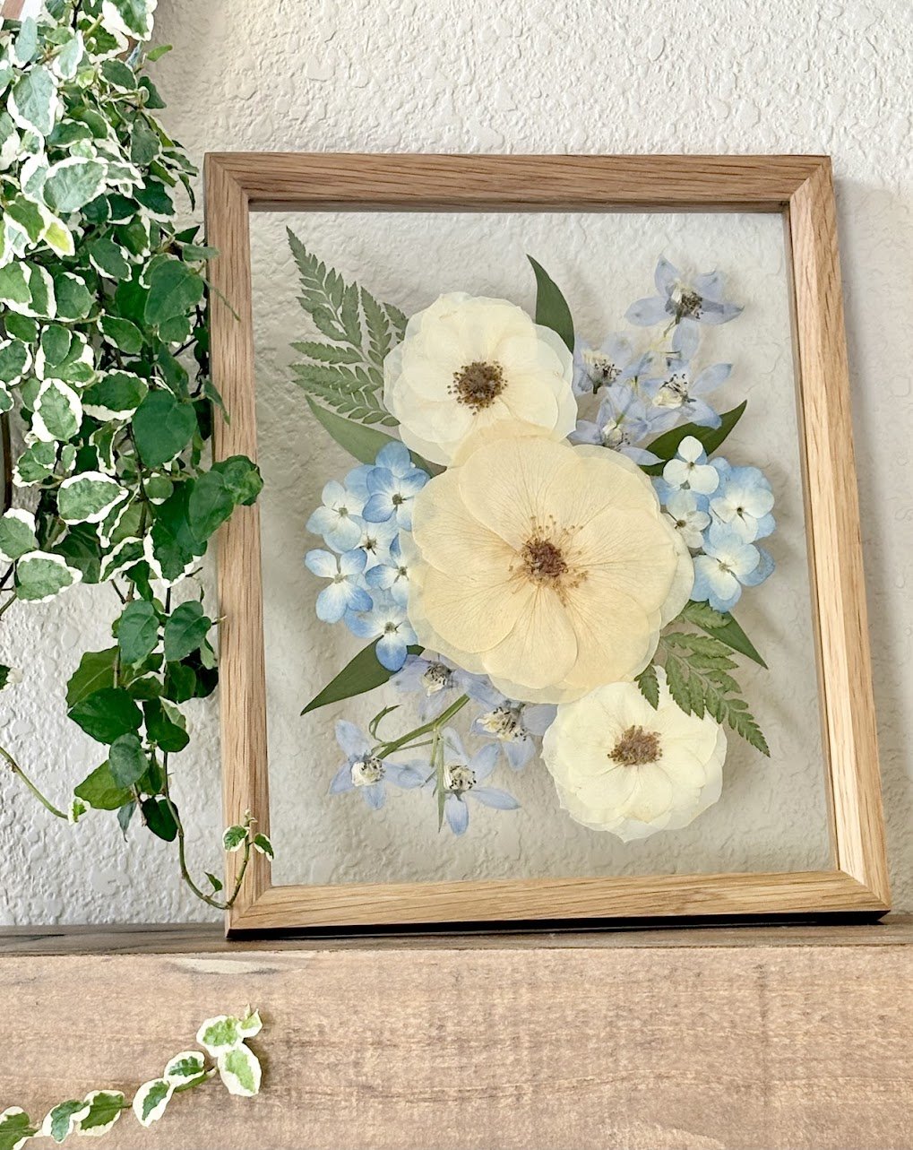 A framed floral arrangement with dried flowers and green leaves on a wall, next to green and white variegated ivy plant.
