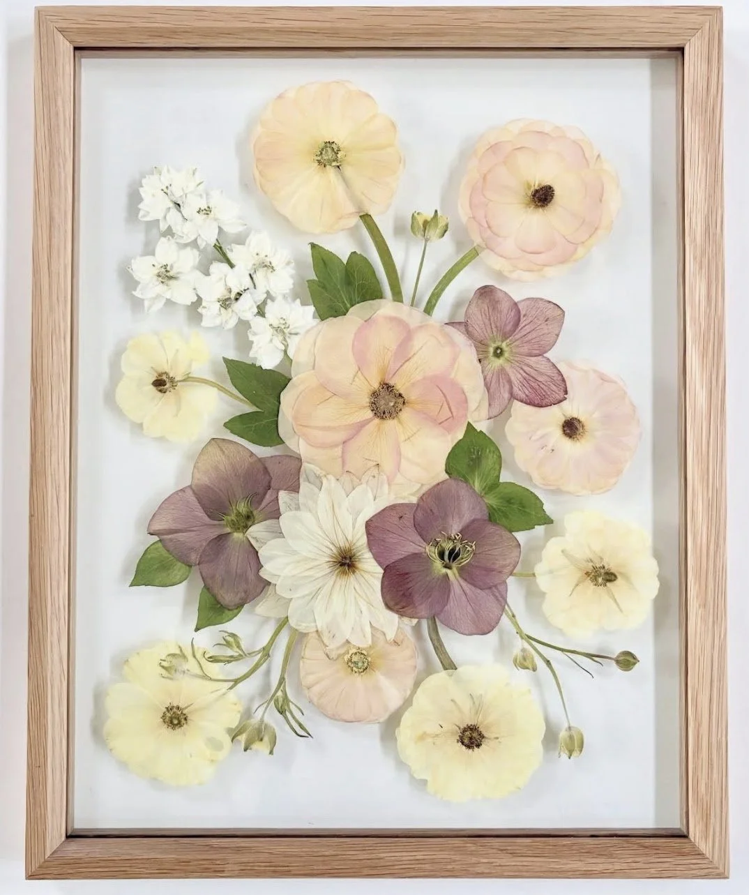 Framed pressed flower arrangement with various pastel-colored and white flowers and green leaves.