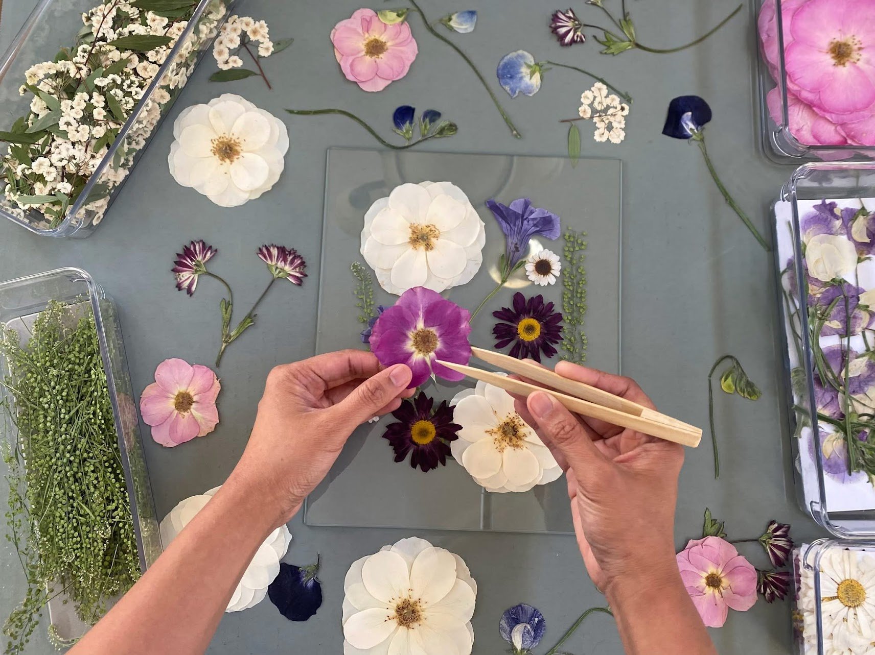 A person arranging purple and white flowers on a glass surface for floral design, with various flowers and leaves scattered around.