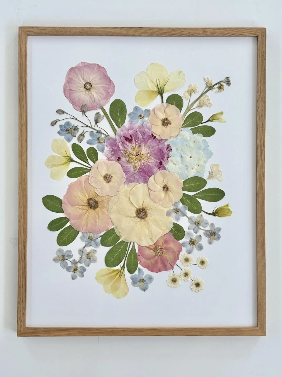 Framed pressed flower art featuring pastel-colored flowers and green leaves on a white background.