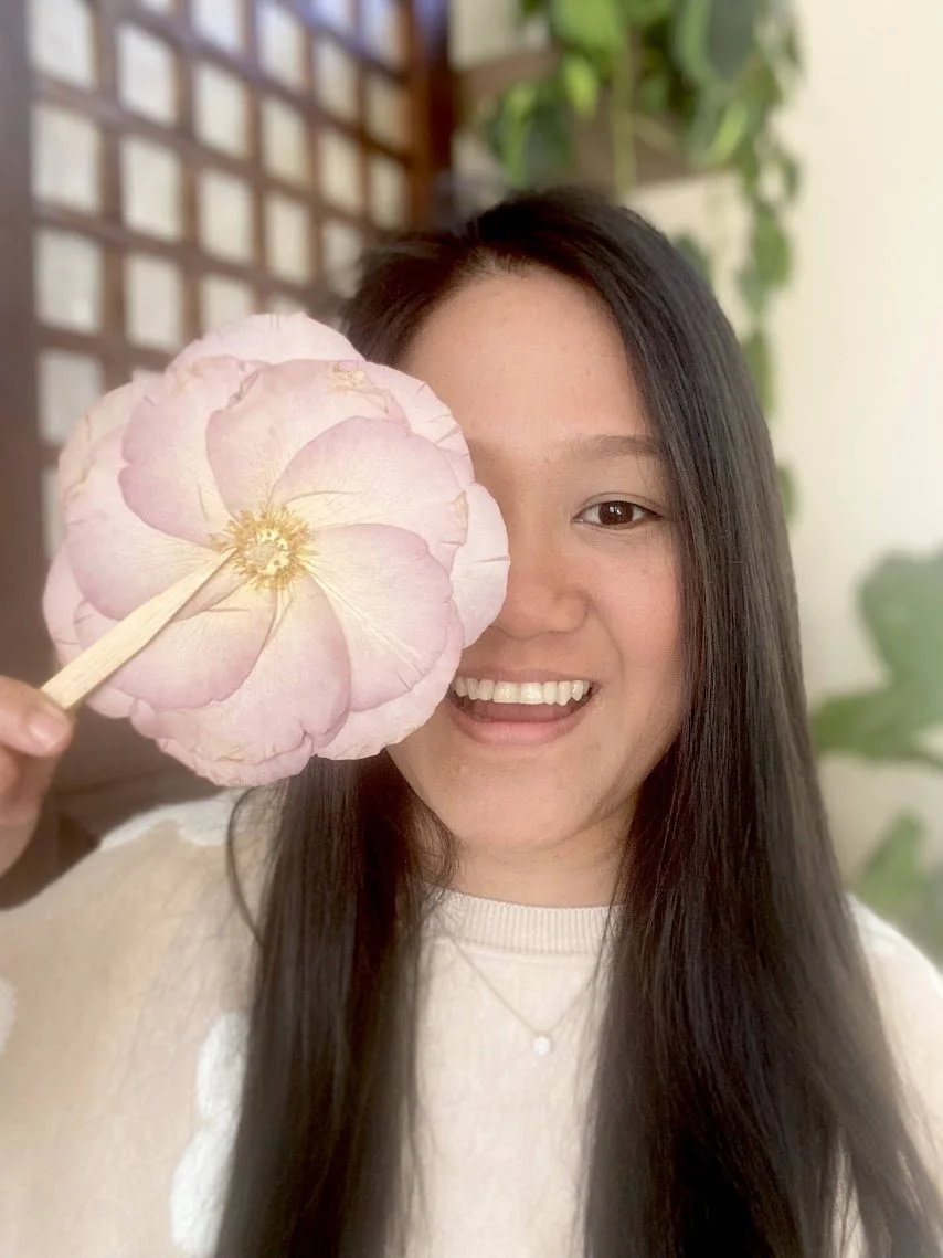 A smiling woman with long dark hair holding a pink flower up to her face, partially covering her eye, indoors with a plant and a wooden grid in the background.